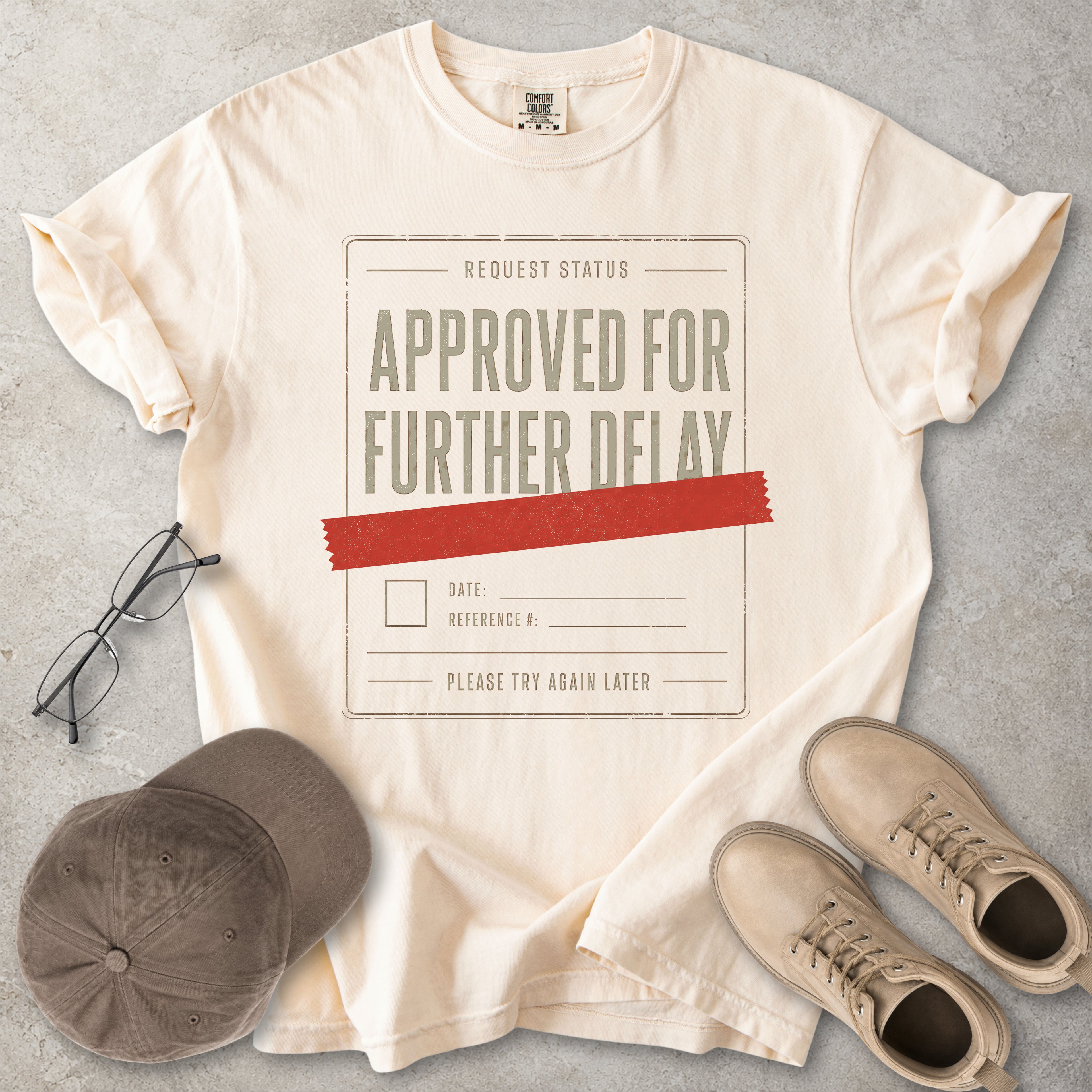 Ivory beige Comfort Colors 1717 T-shirt with a funny Approved for Further Delay graphic showing a request status form covered with red tape.