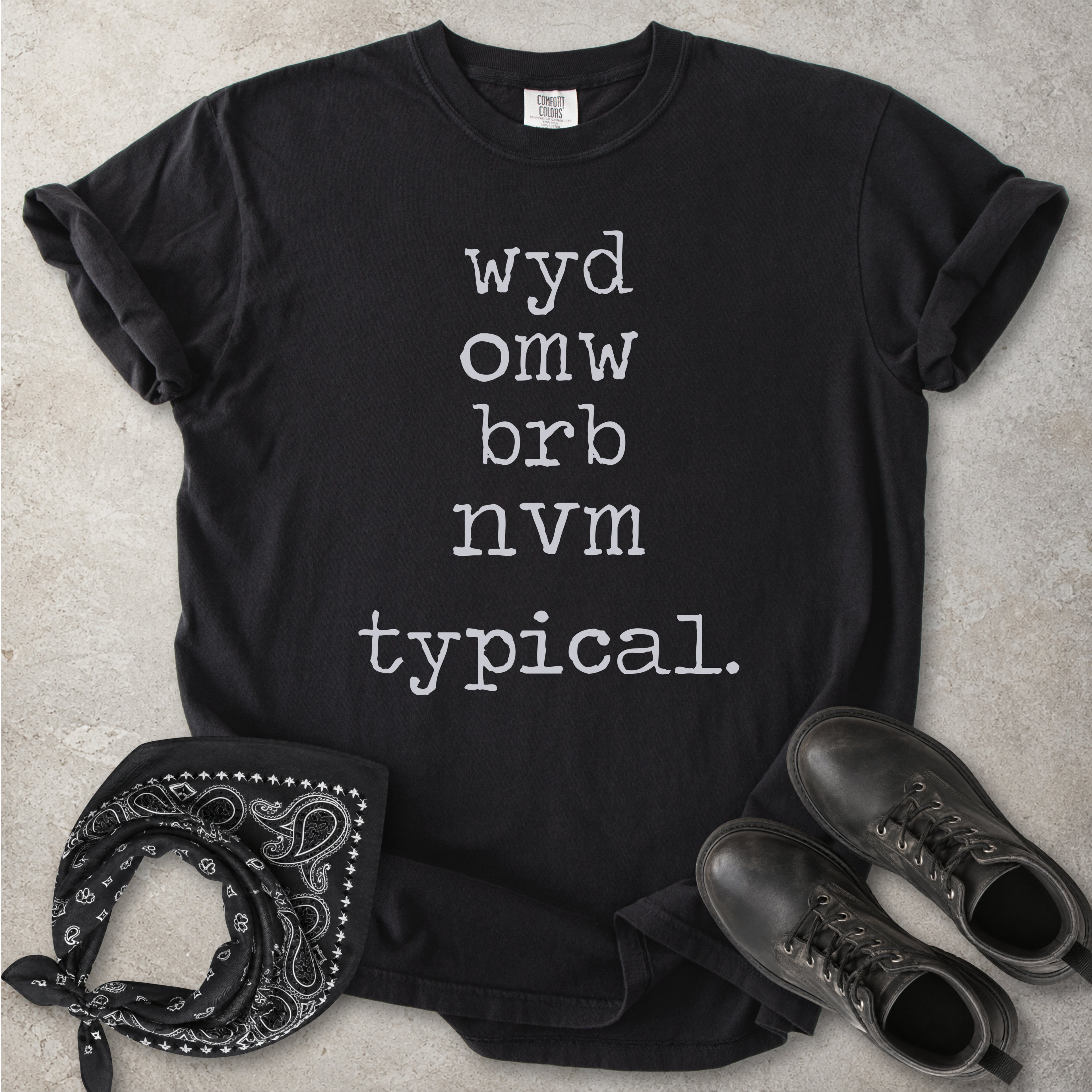 Black Comfort Colors graphic T-shirt with black text reading wyd omw brb nvm typical, styled with black boots and a black bandana