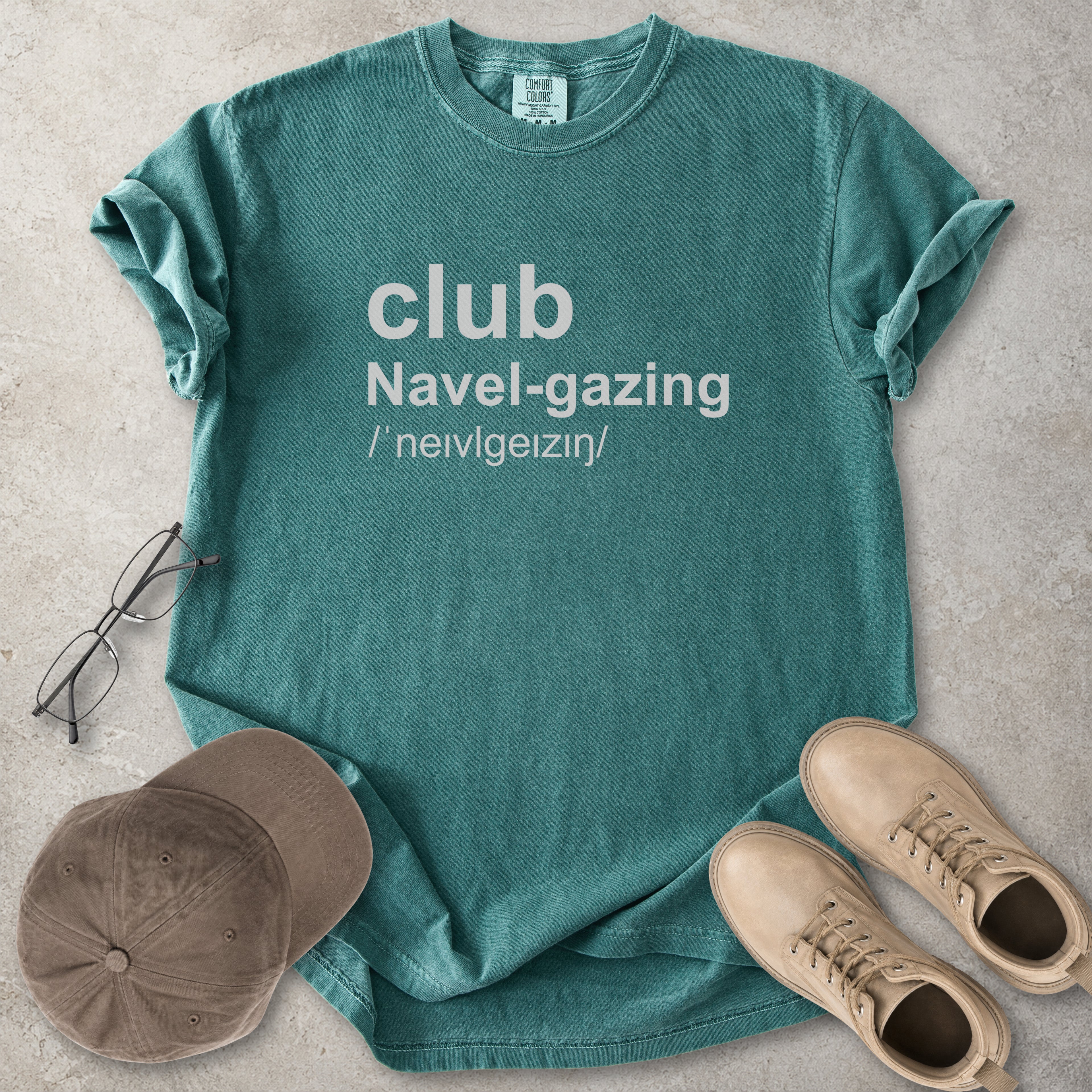 Blue spruce Comfort Colors graphic T-shirt with grey text reading club navel-gazing, styled with notebook, pen, glasses and tan boots