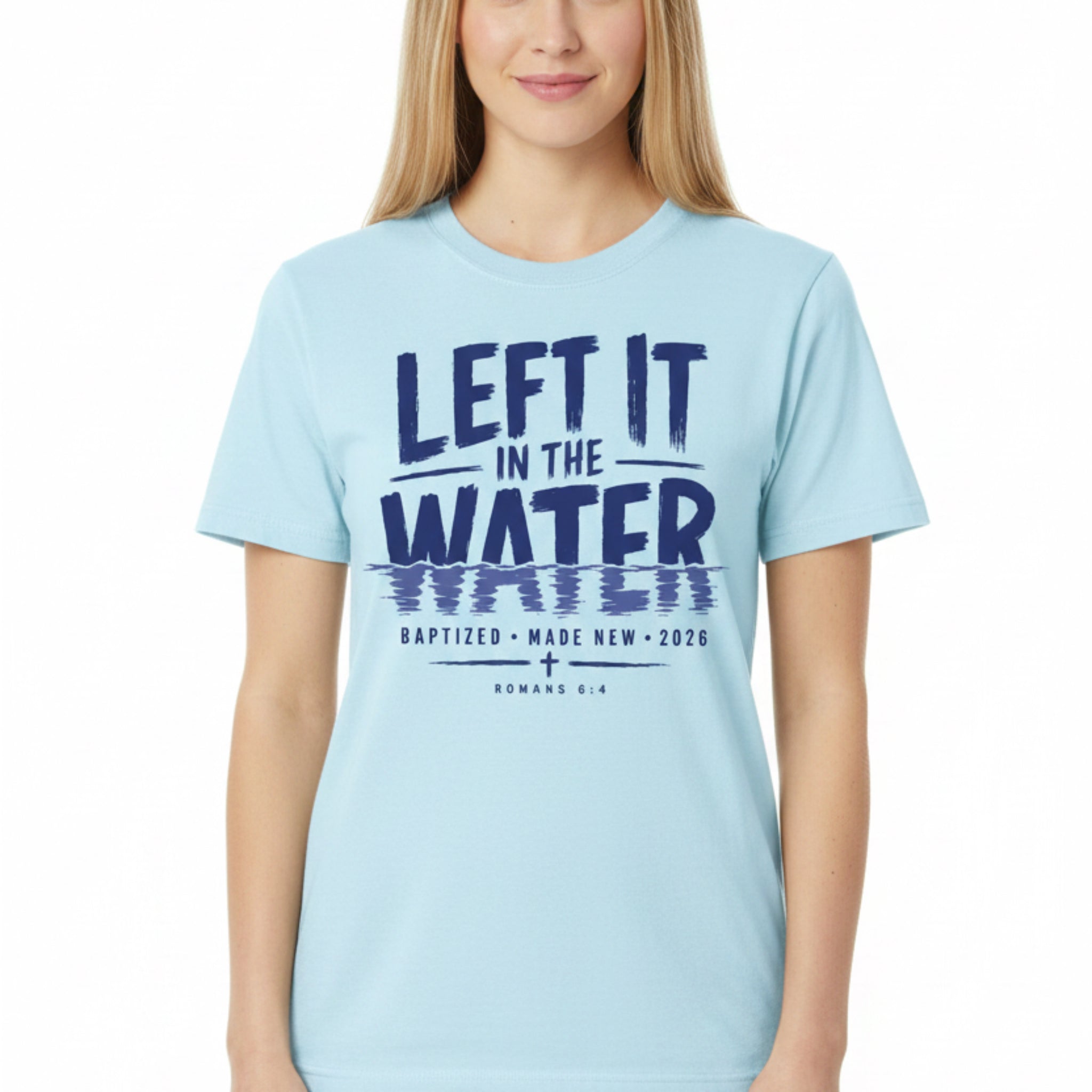 A light blue short-sleeve t-shirt laid flat on a rustic dark wood background, featuring a dark navy blue graphic print that reads 'LEFT IT IN THE WATER' with a reflection effect, and smaller text below reading 'BAPTIZED MADE NEW 2026 ROMANS 6:4'.