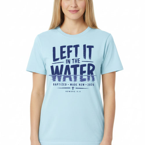 A light blue short-sleeve t-shirt laid flat on a rustic dark wood background, featuring a dark navy blue graphic print that reads 'LEFT IT IN THE WATER' with a reflection effect, and smaller text below reading 'BAPTIZED MADE NEW 2026 ROMANS 6:4'.