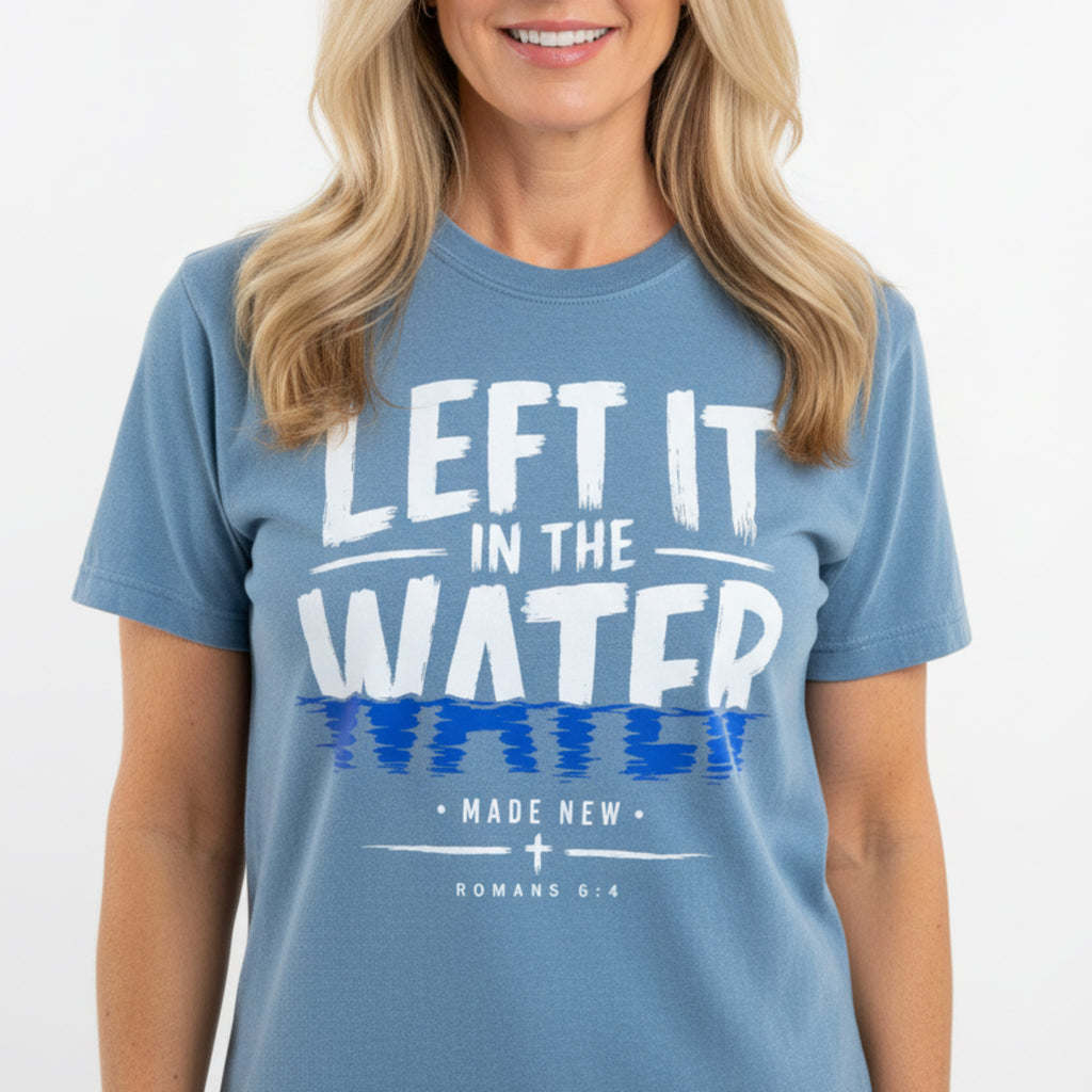 A heathered blue short-sleeve t-shirt with a graphic print reading 'LEFT IT IN THE WATER' in white and blue, with 'MADE NEW ROMANS 6:4' below, laid flat on a rustic wooden background.