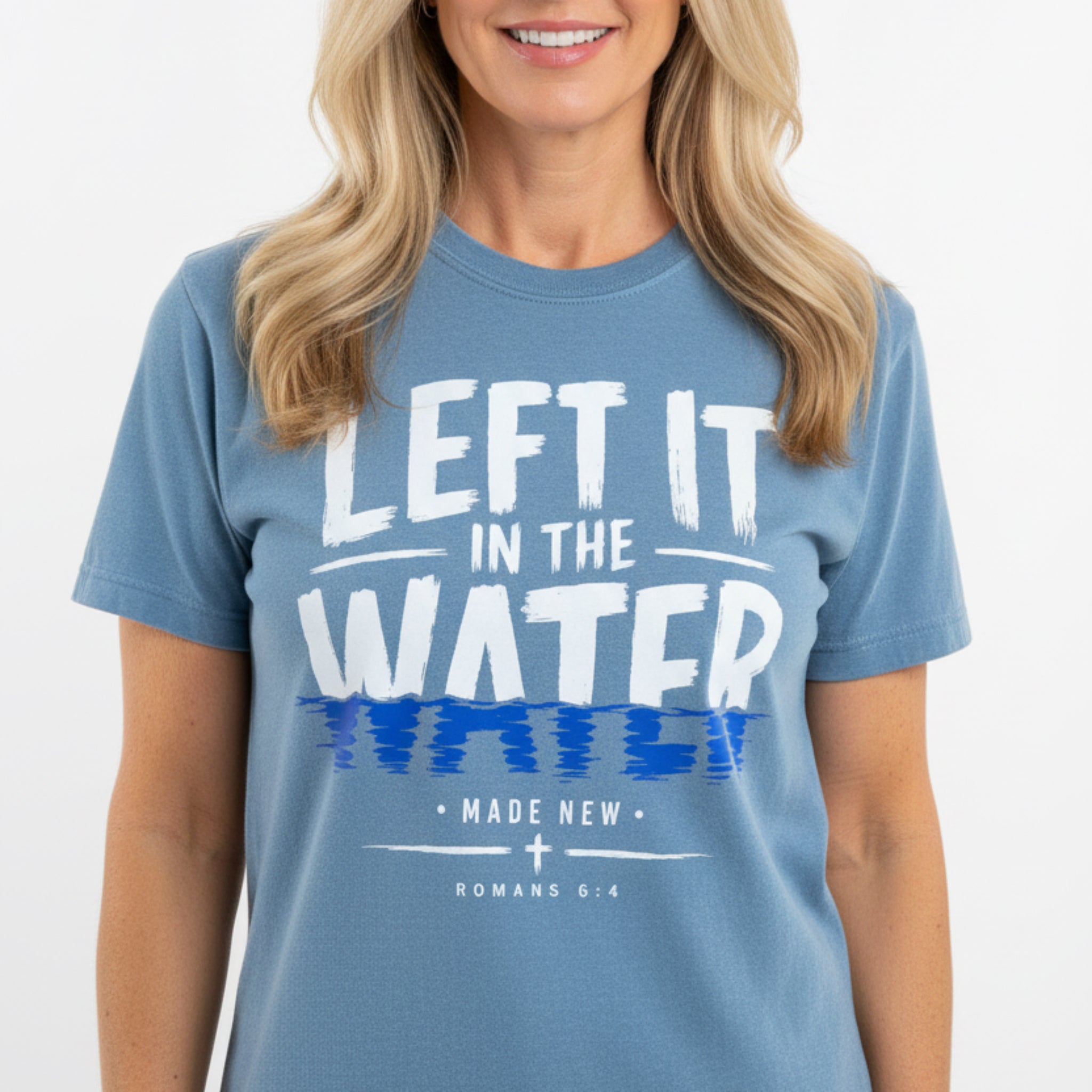 A heathered blue short-sleeve t-shirt with a graphic print reading 'LEFT IT IN THE WATER' in white and blue, with 'MADE NEW ROMANS 6:4' below, laid flat on a rustic wooden background.