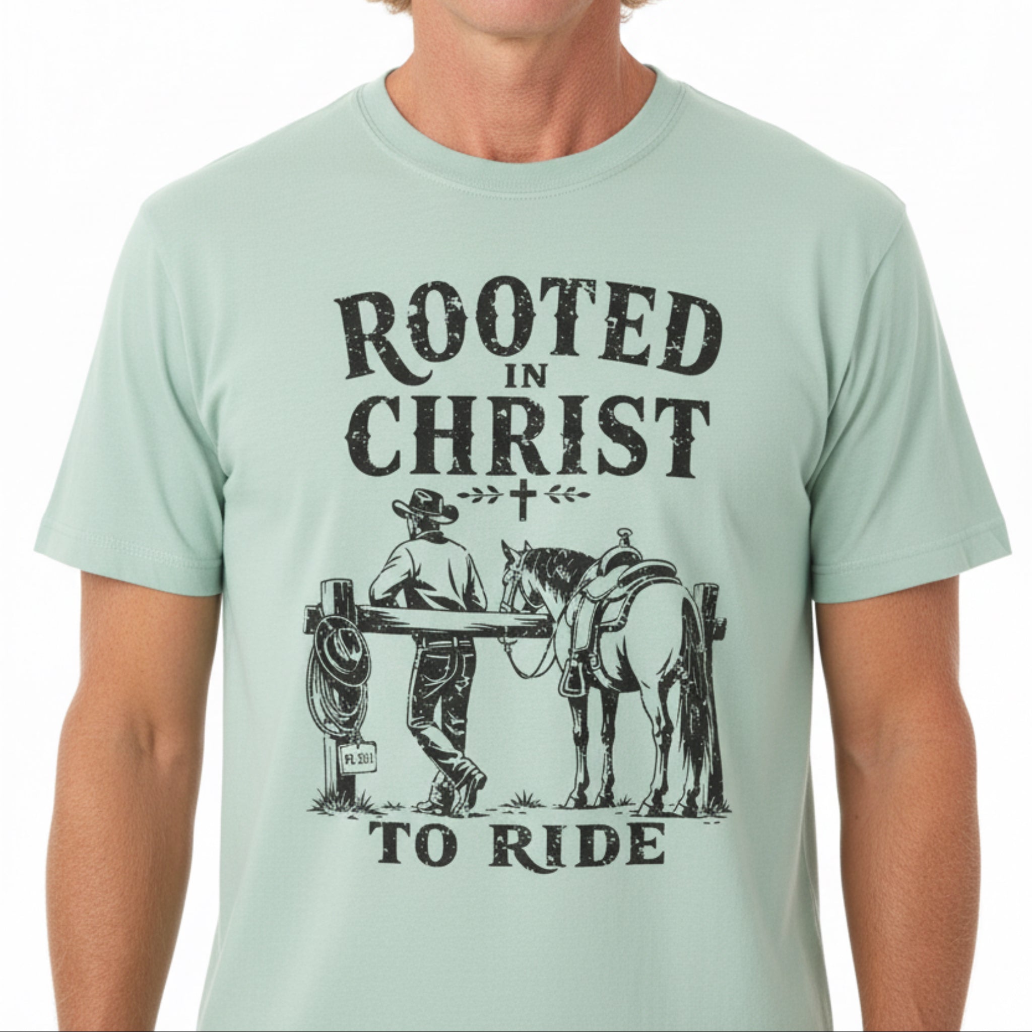 A light mint green t-shirt with a black graphic print reading 'ROOTED IN CHRIST TO RIDE' and showing an illustration of a cowboy standing by a horse at a fence, laid flat on a rustic wooden background.