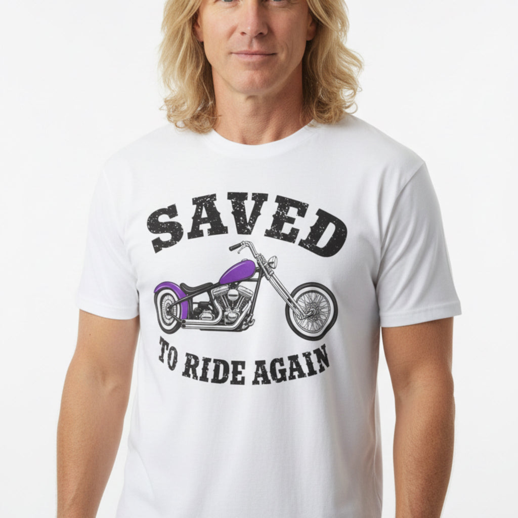 A white short-sleeve graphic T-shirt laid flat on a rustic wooden surface. The shirt features a distressed black text design reading 'SAVED TO RIDE AGAIN' surrounding an illustration of a chopper motorcycle with a purple fuel tank.