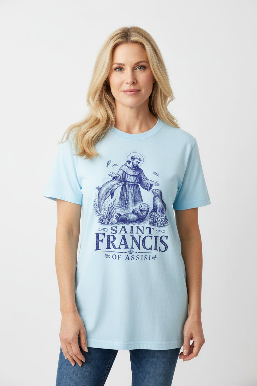 A light blue short-sleeve t-shirt laid flat on a rustic wooden background, featuring a dark blue graphic of Saint Francis with a dolphin and otters, and the text 'SAINT FRANCIS OF ASSISI'.