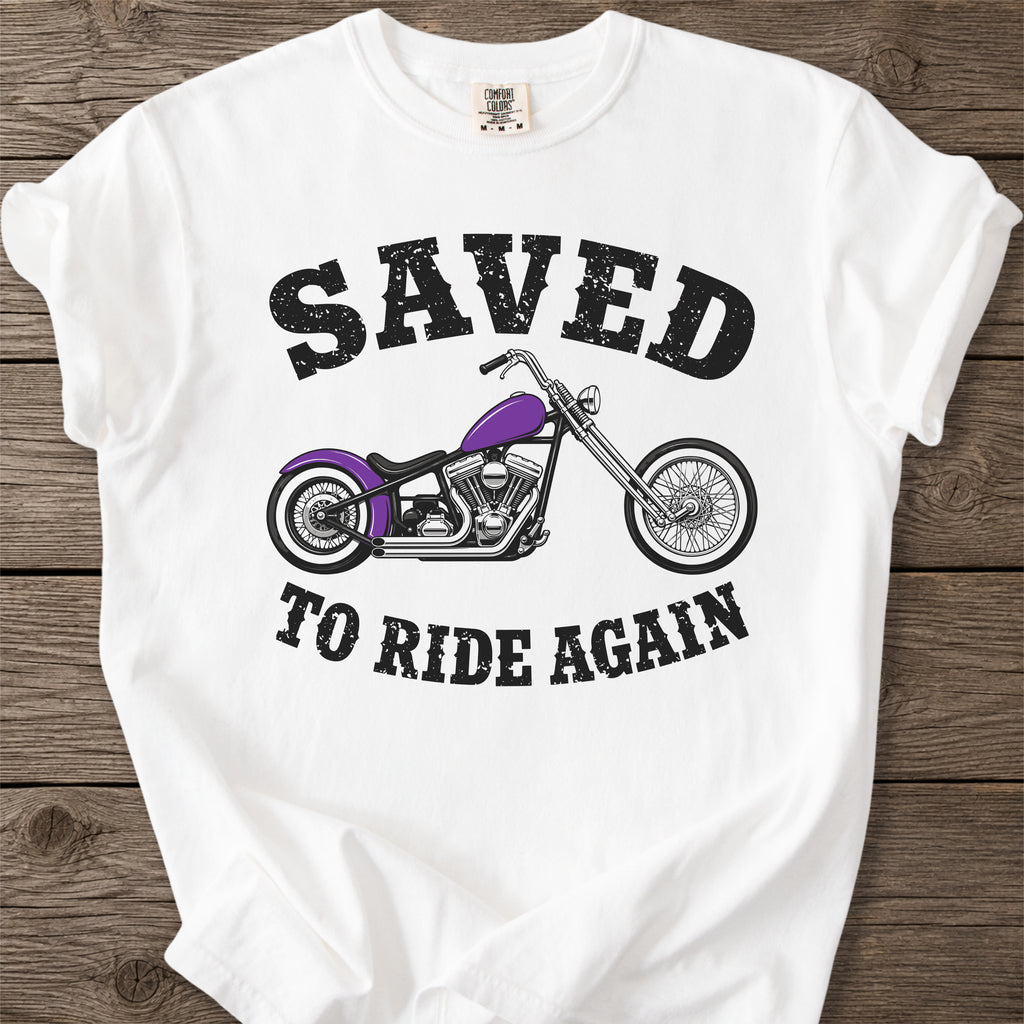 A white short-sleeve graphic T-shirt laid flat on a rustic wooden surface. The shirt features a distressed black text design reading 'SAVED TO RIDE AGAIN' surrounding an illustration of a chopper motorcycle with a purple fuel tank.