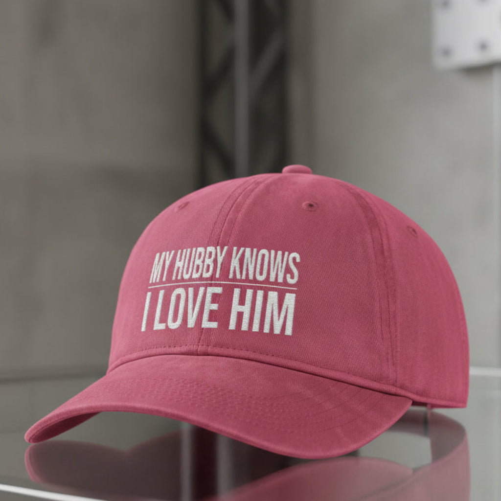 A dusty rose pink baseball cap with white embroidered text reading 'MY HUBBY KNOWS I LOVE HIM', resting on a reflective glass surface against a gray concrete wall background.