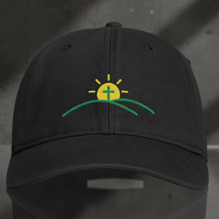 A black baseball cap featuring an embroidered logo on the front with a yellow sun, green hills, and a white cross, set against a plain white background.