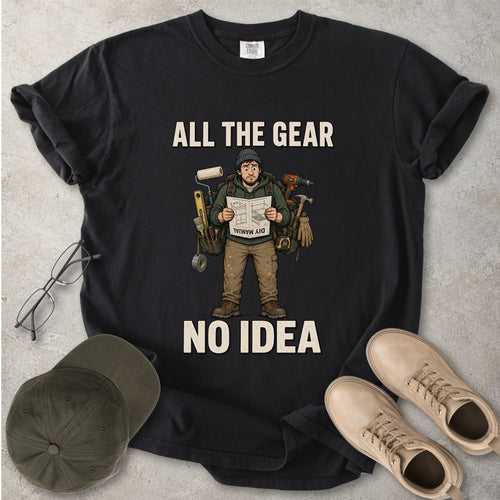 Black t-shirt with graphic design and text 'All the Gear No Idea' on a gray surface with a cap and boots.