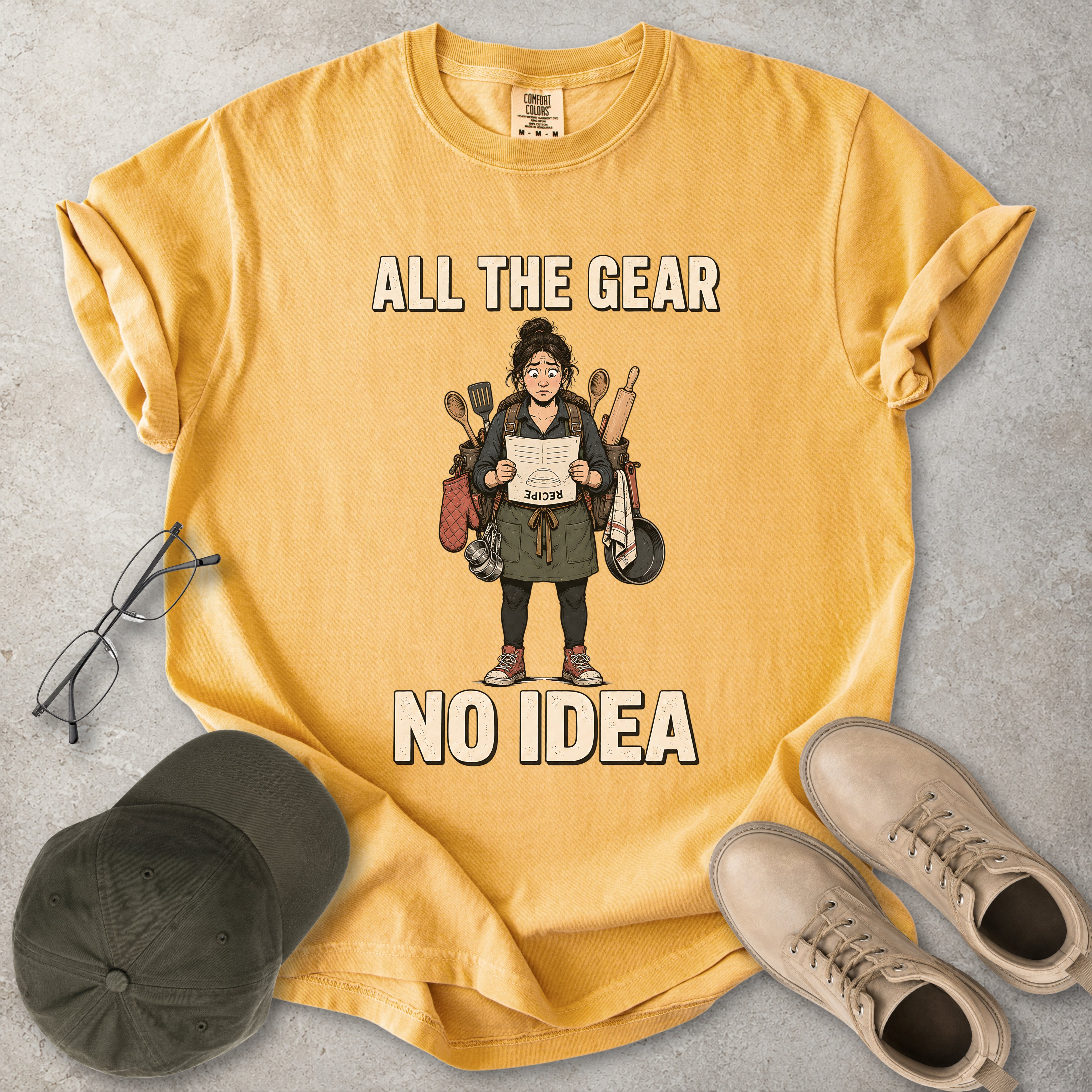 Mustard yellow Comfort Colors 1717 T-shirt with a funny All the Gear No Idea cooking graphic showing a woman holding an upside-down recipe with kitchen utensils.