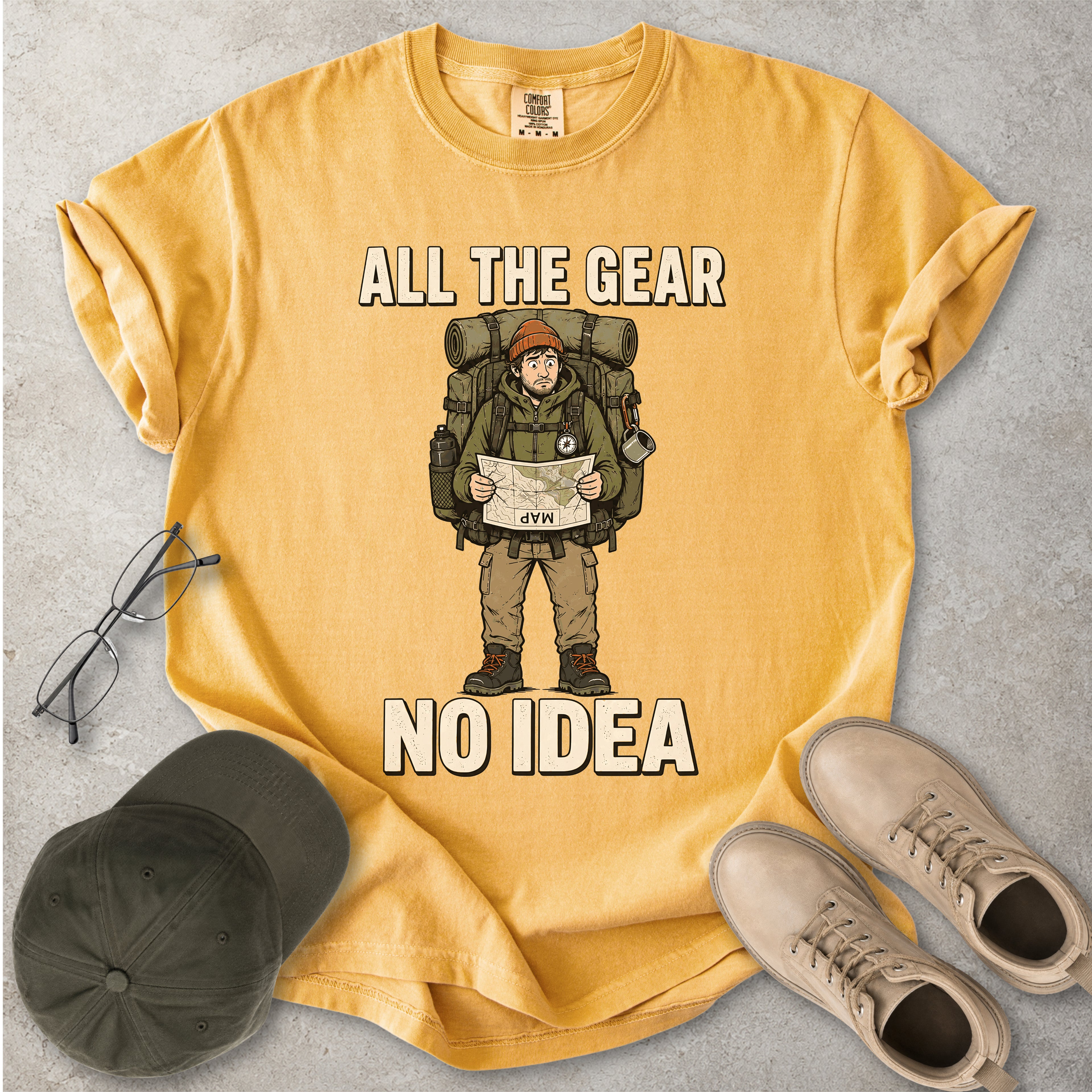 Mustard yellow Comfort Colors 1717 T-shirt with a funny All the Gear No Idea hiking graphic showing a confused hiker holding an upside-down map with a large backpack.