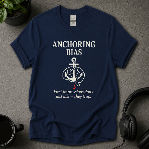 Navy blue t-shirt with 'Anchoring Bias' graphic and text on a gray surface with a plant and headphones.