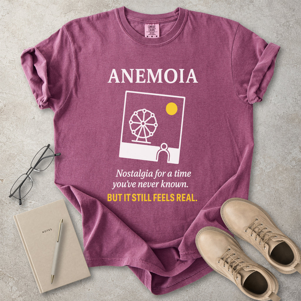 Purple t-shirt with 'ANEMOIA' design and text, placed on a concrete surface with a notebook, pen, and shoes.