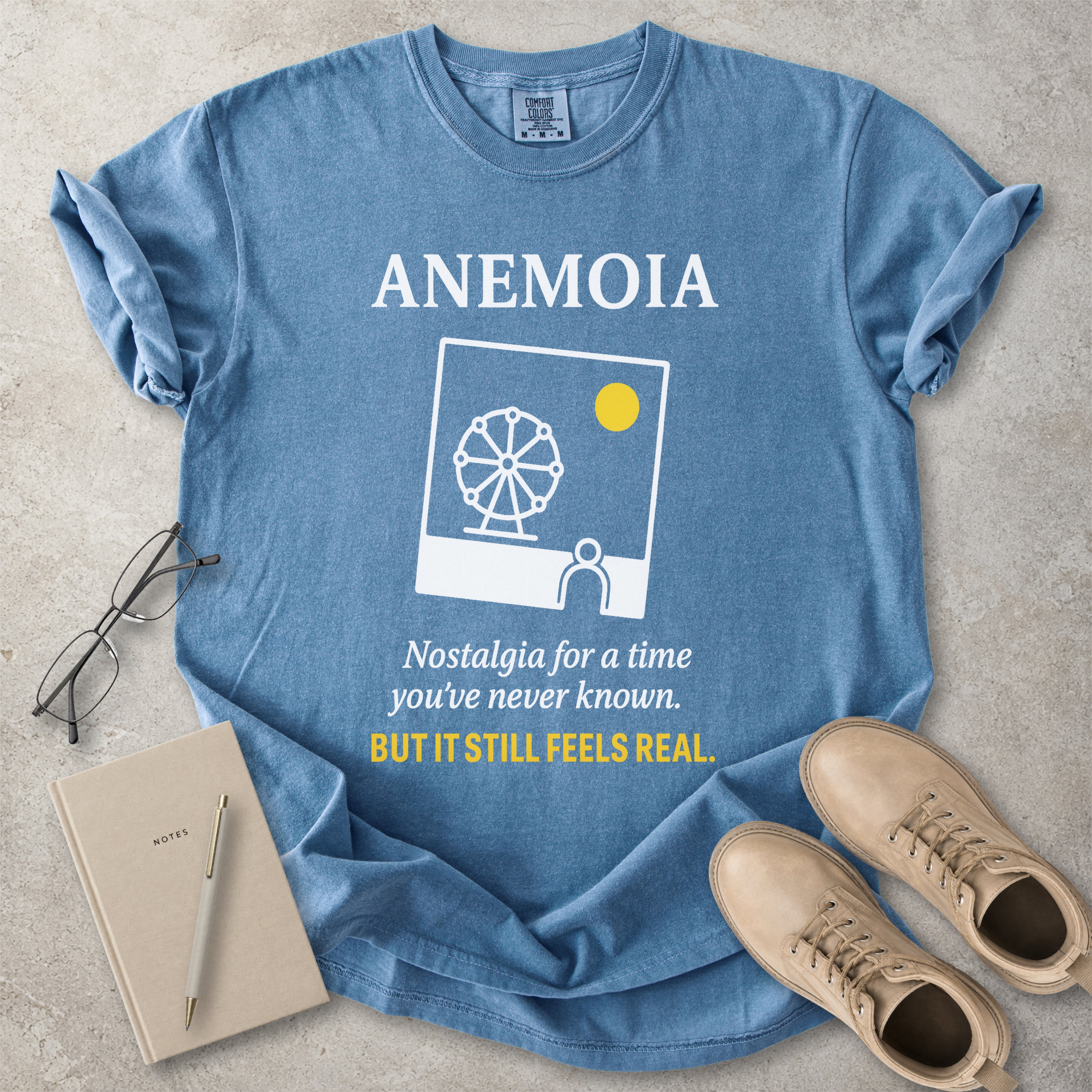 Blue t-shirt with 'Anemoia' design and text, placed on a concrete surface with a notebook, pen, and shoes.