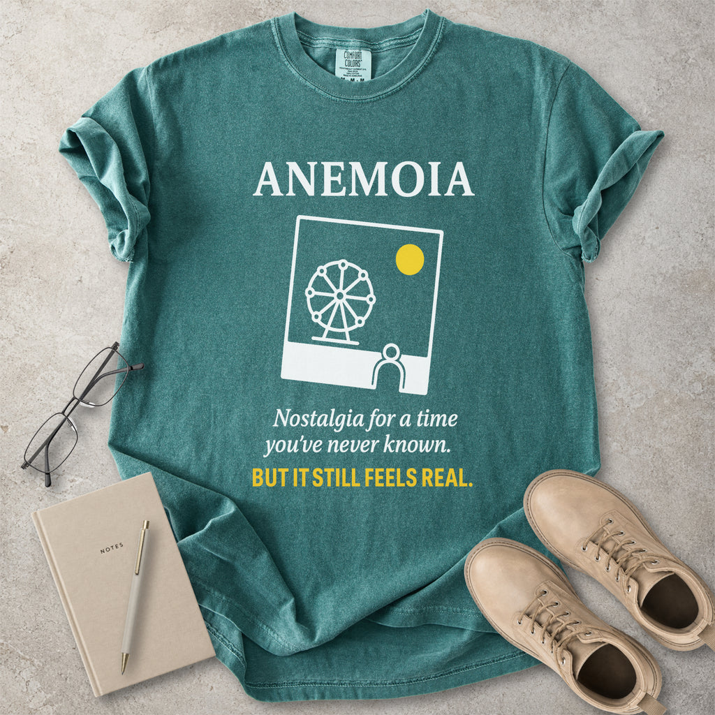 Green t-shirt with 'ANEMOIA' design and text, placed on a concrete surface with a notebook, pen, and shoes.