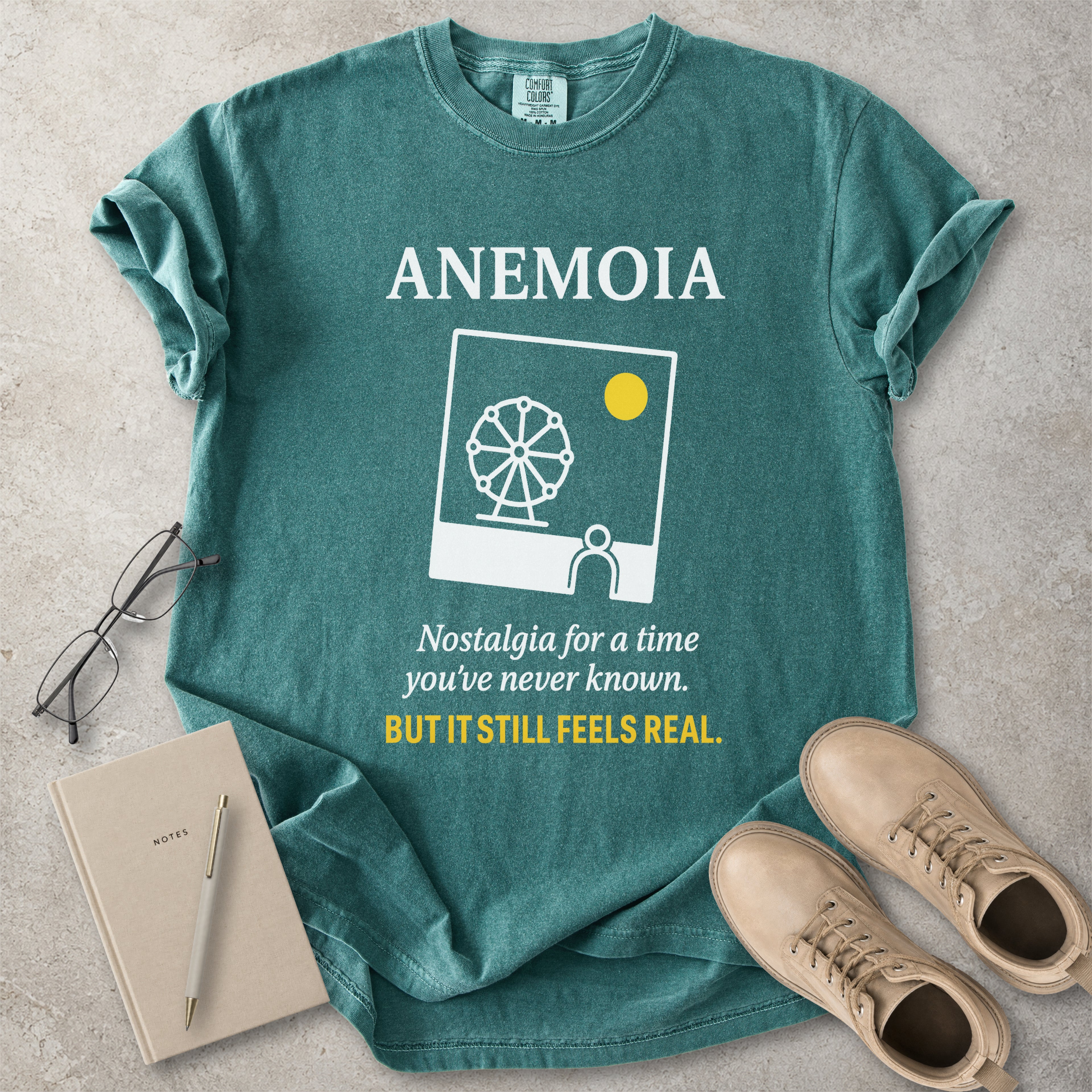 Green t-shirt with 'ANEMOIA' design and text, placed on a concrete surface with a notebook, pen, and shoes.
