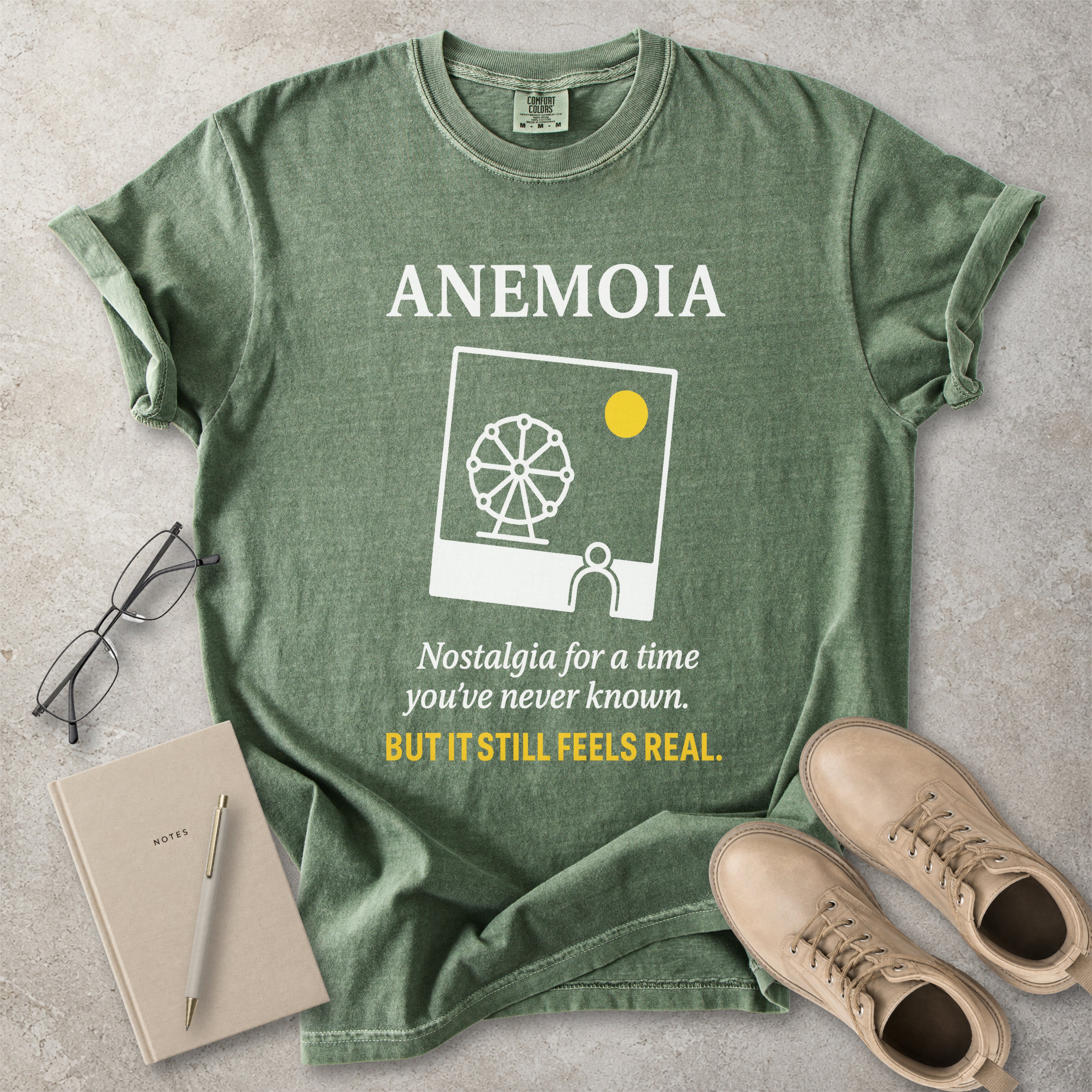 Green t-shirt with 'Anemoia' design and text on a concrete surface with shoes and notebook.
