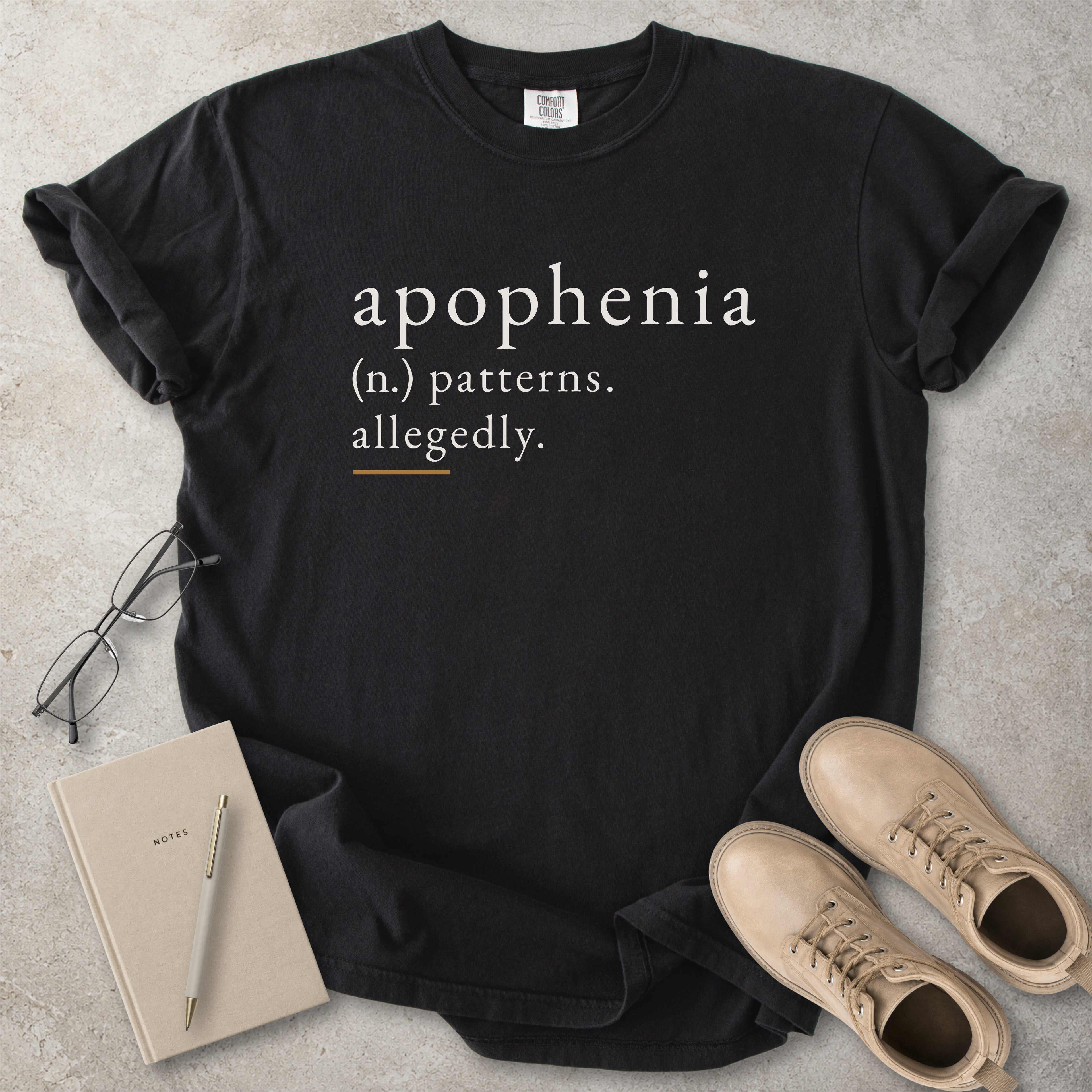 Black Comfort Colors graphic T-shirt with apophenia definition text reading patterns allegedly, styled with notebook, pen, glasses and tan boots