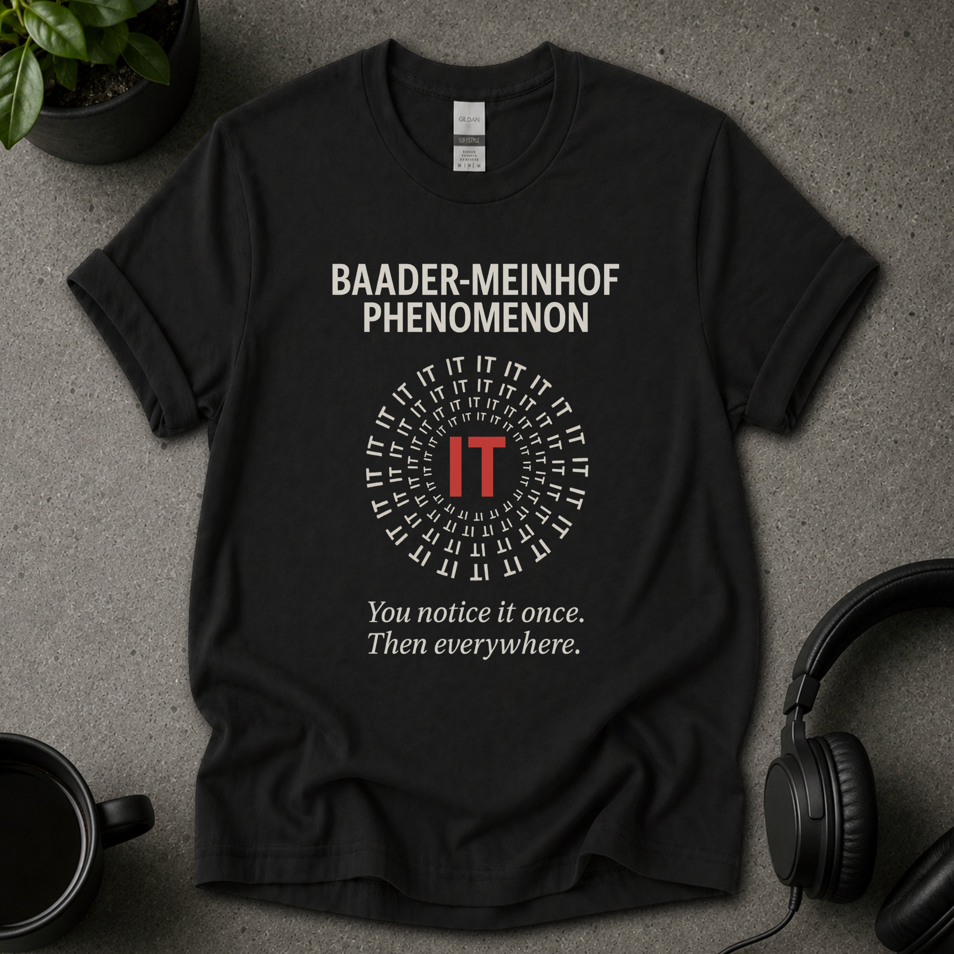 Black t-shirt with 'Baader-Meinhof Phenomenon' text on a gray surface with headphones and a mug.