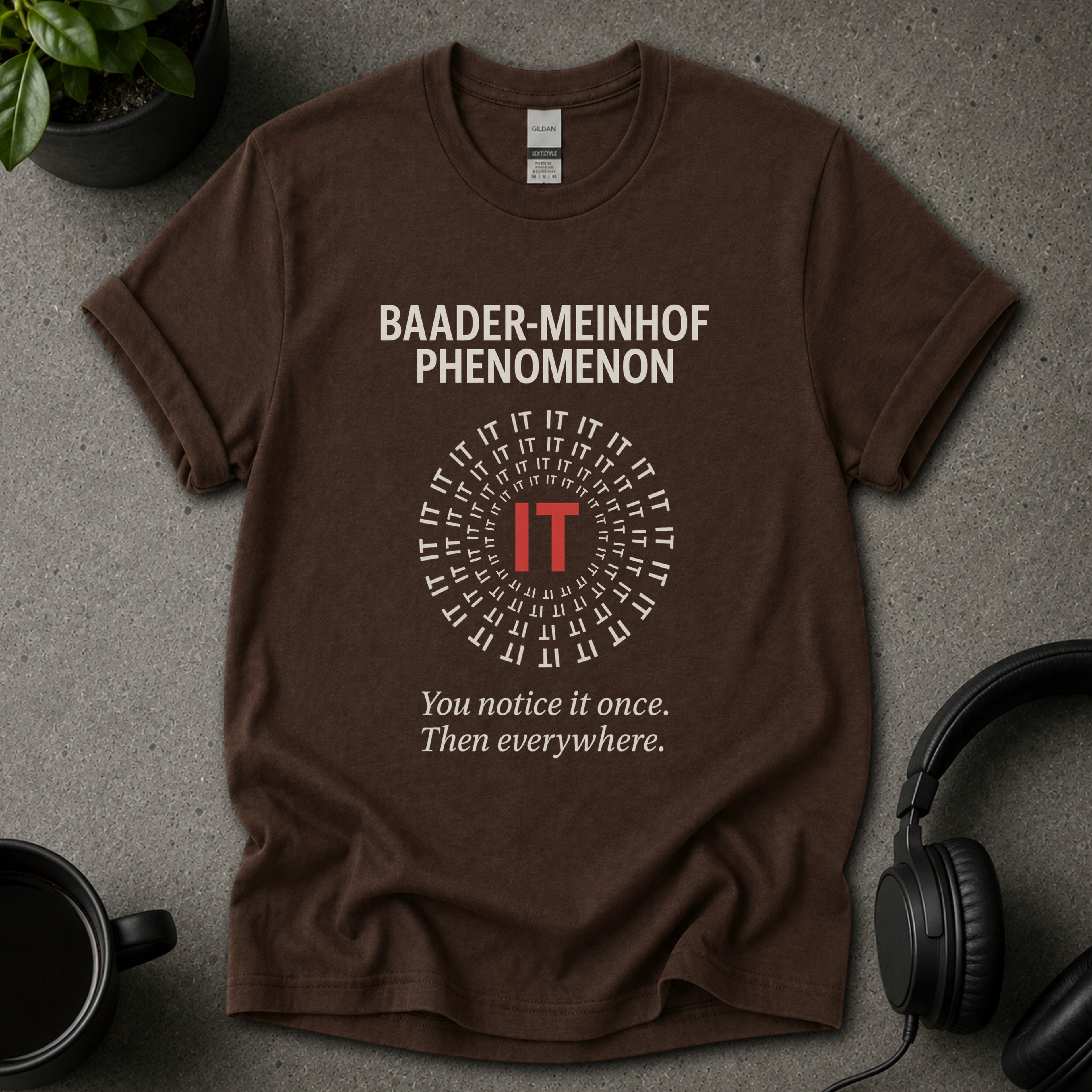 Brown t-shirt with 'Baader-Meinhof Phenomenon' text on a gray surface with headphones and a mug.