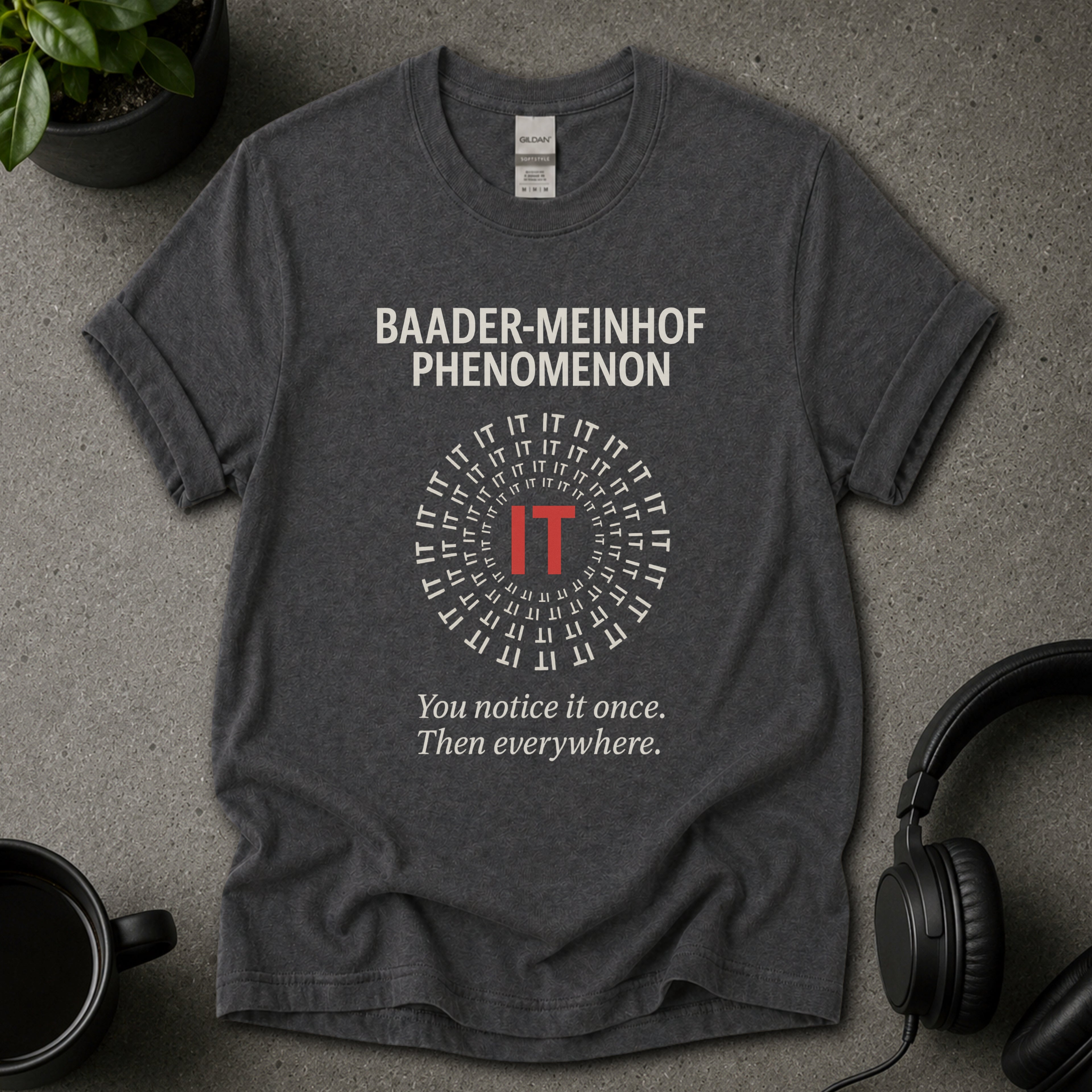 Grey t-shirt with 'Baader-Meinhof Phenomenon' text on a gray surface with headphones and a mug.