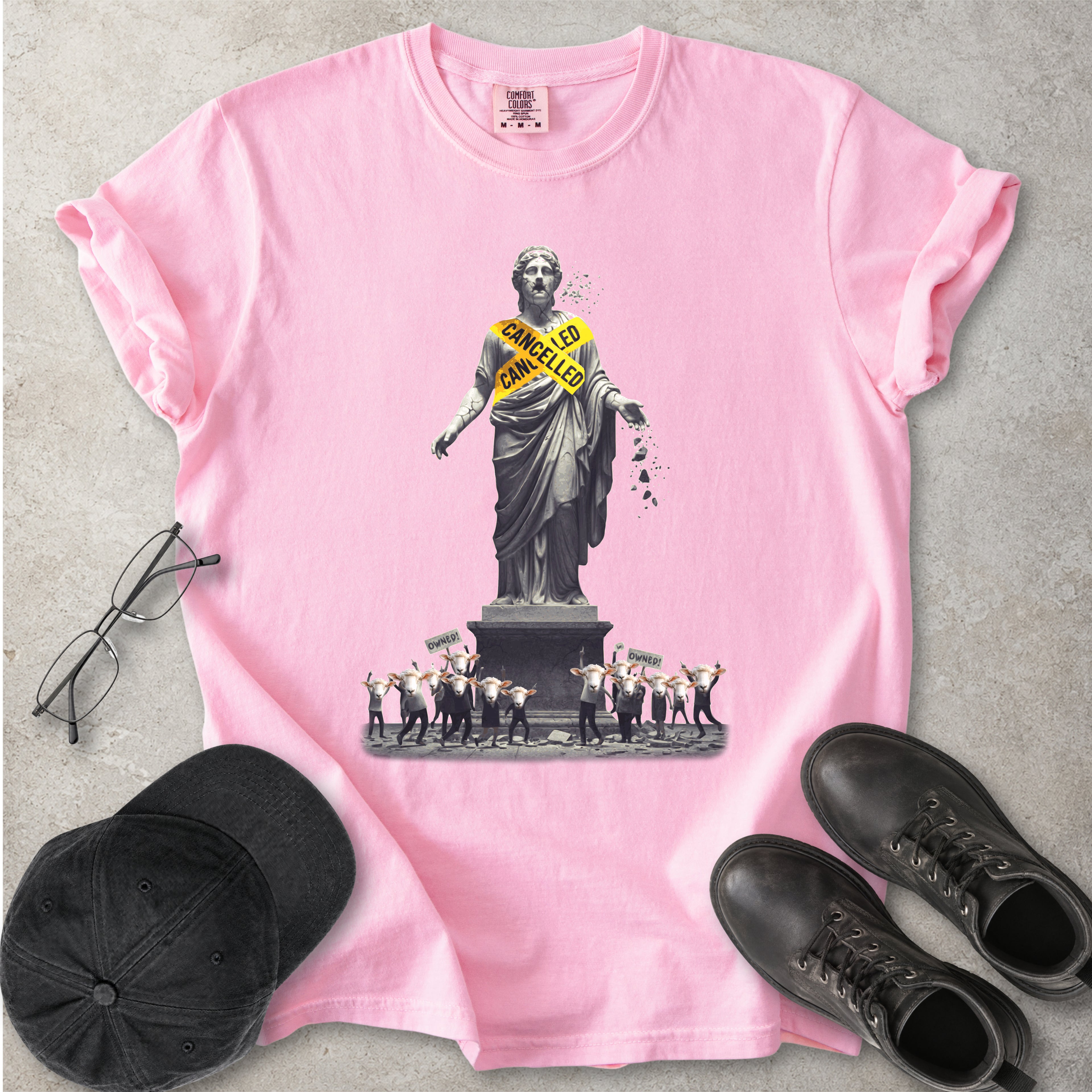 Pink Comfort Colors graphic T-shirt with cancelled statue and crowd design, styled with black cap, glasses and black boots