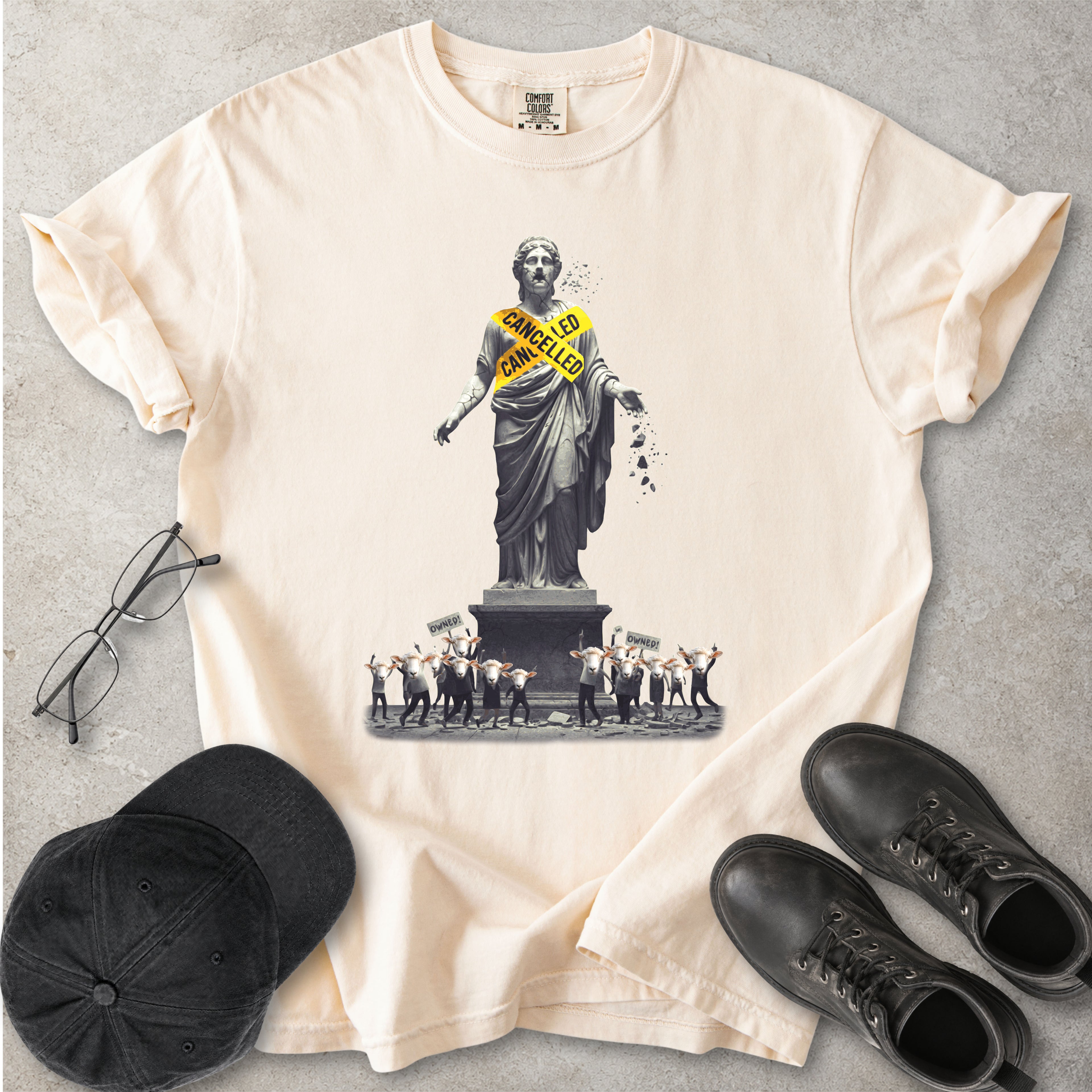 Beige Comfort Colors graphic T-shirt with cancelled statue and crowd design, styled with black cap, glasses and black boots