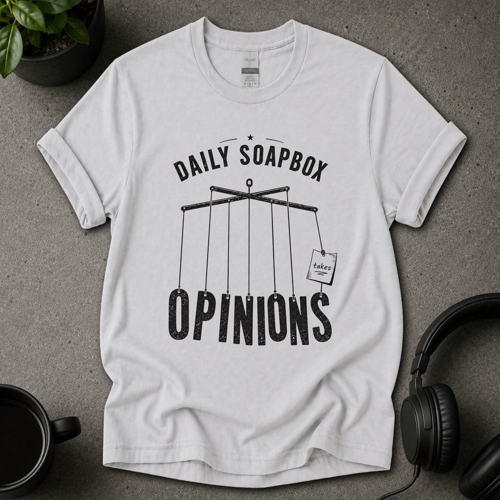 Ice grey t-shirt with 'Daily Soapbox Opinions' graphic on a gray surface with a plant and mug in the background.