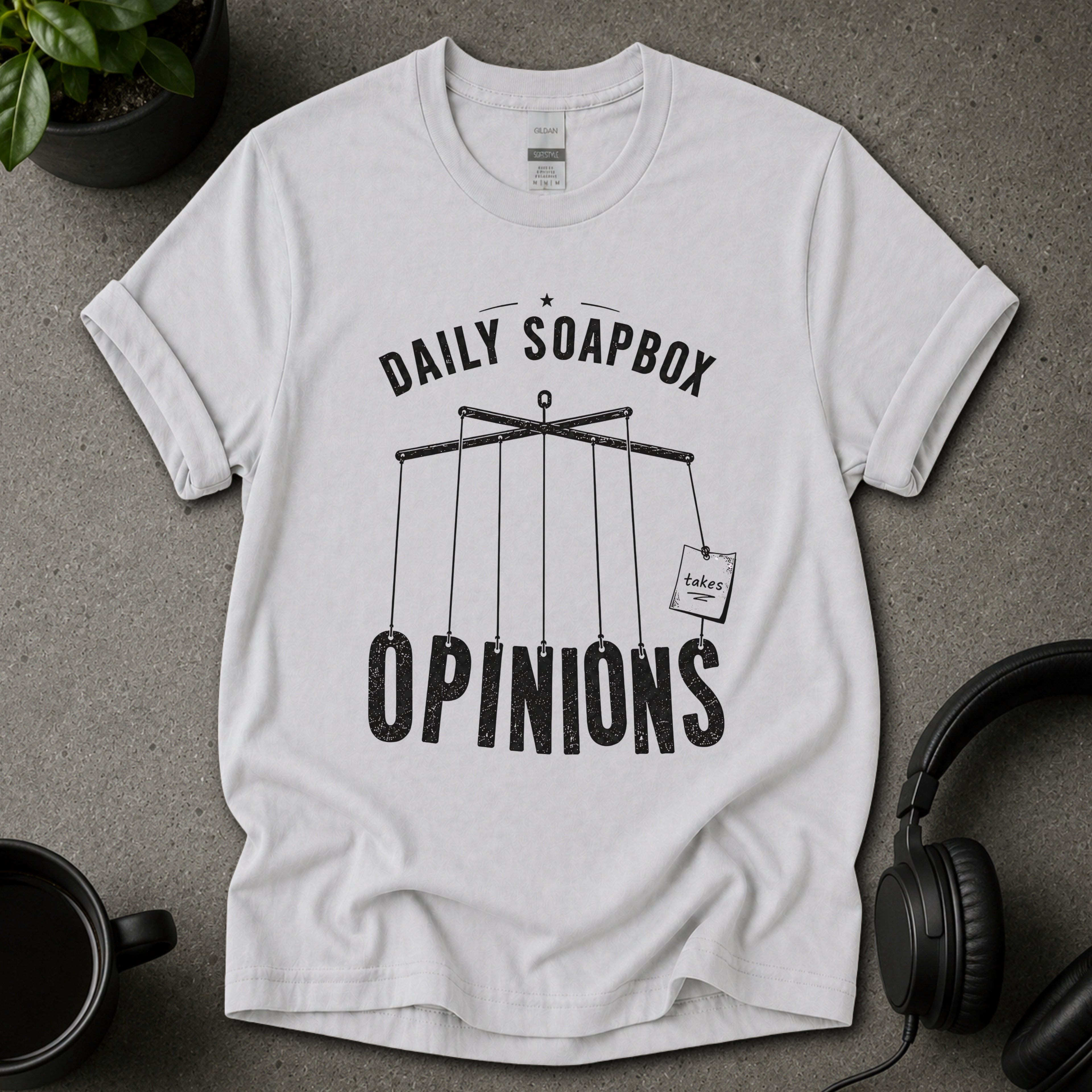 Ice grey t-shirt with 'Daily Soapbox Opinions' graphic on a gray surface with a plant and mug in the background.