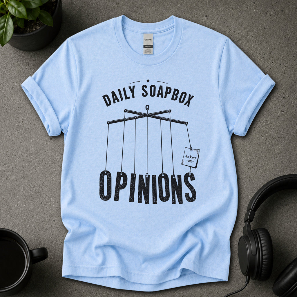 Light blue t-shirt with 'Daily Soapbox Opinions' text on a gray surface with a plant, mug, and headphones.