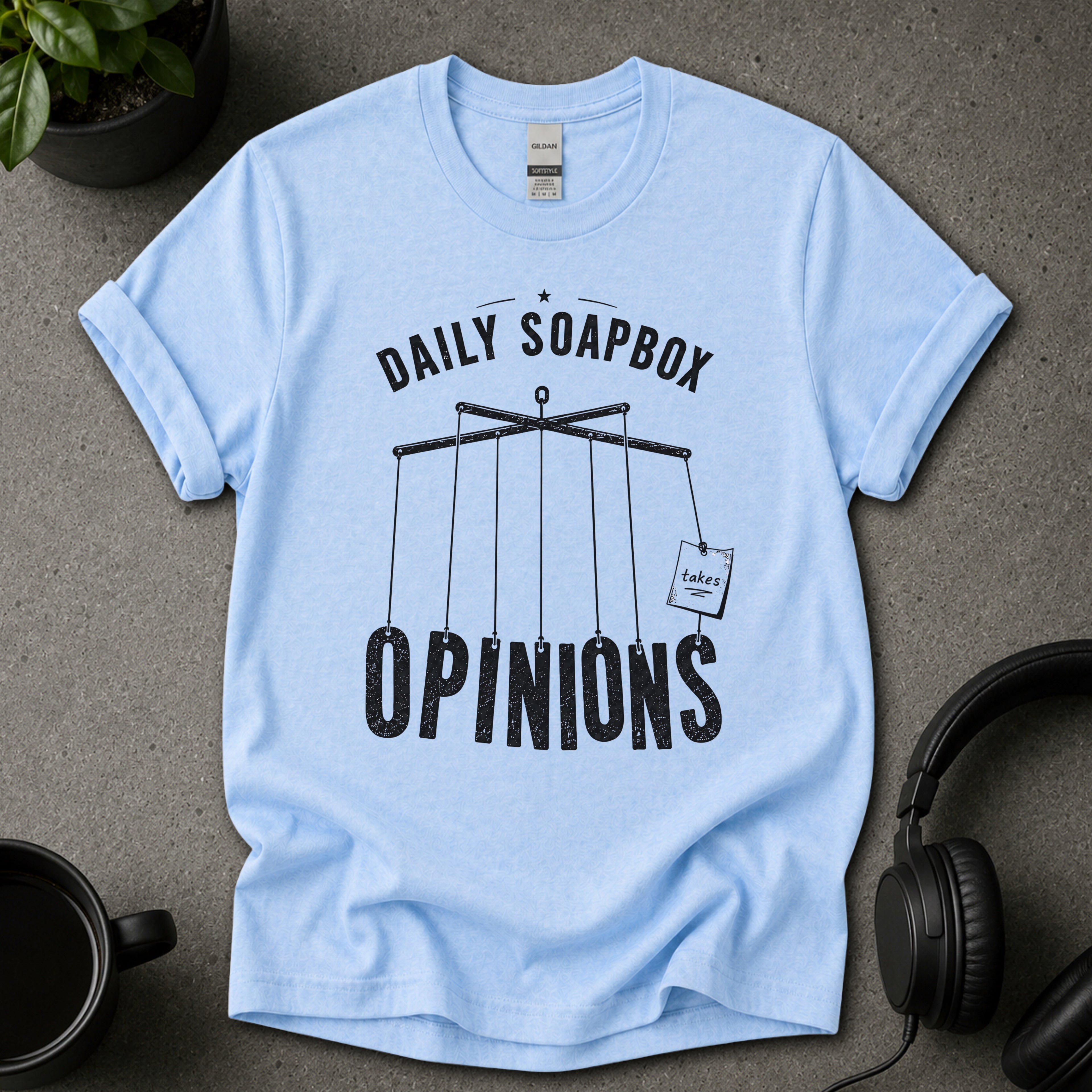 Light blue t-shirt with 'Daily Soapbox Opinions' text on a gray surface with a plant, mug, and headphones.