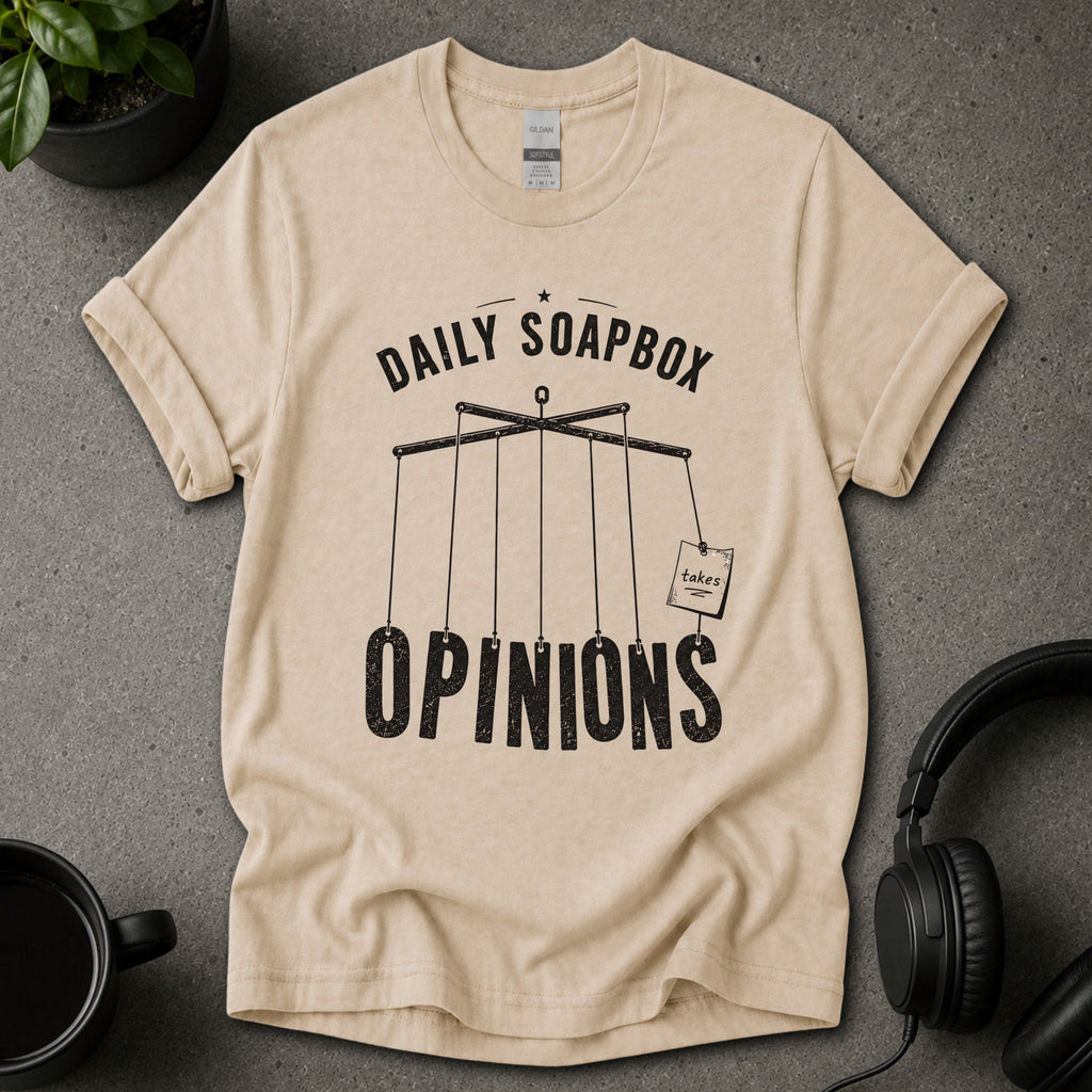 Beige t-shirt with 'Daily Soapbox Opinions' graphic on a gray surface with a mug and headphones.