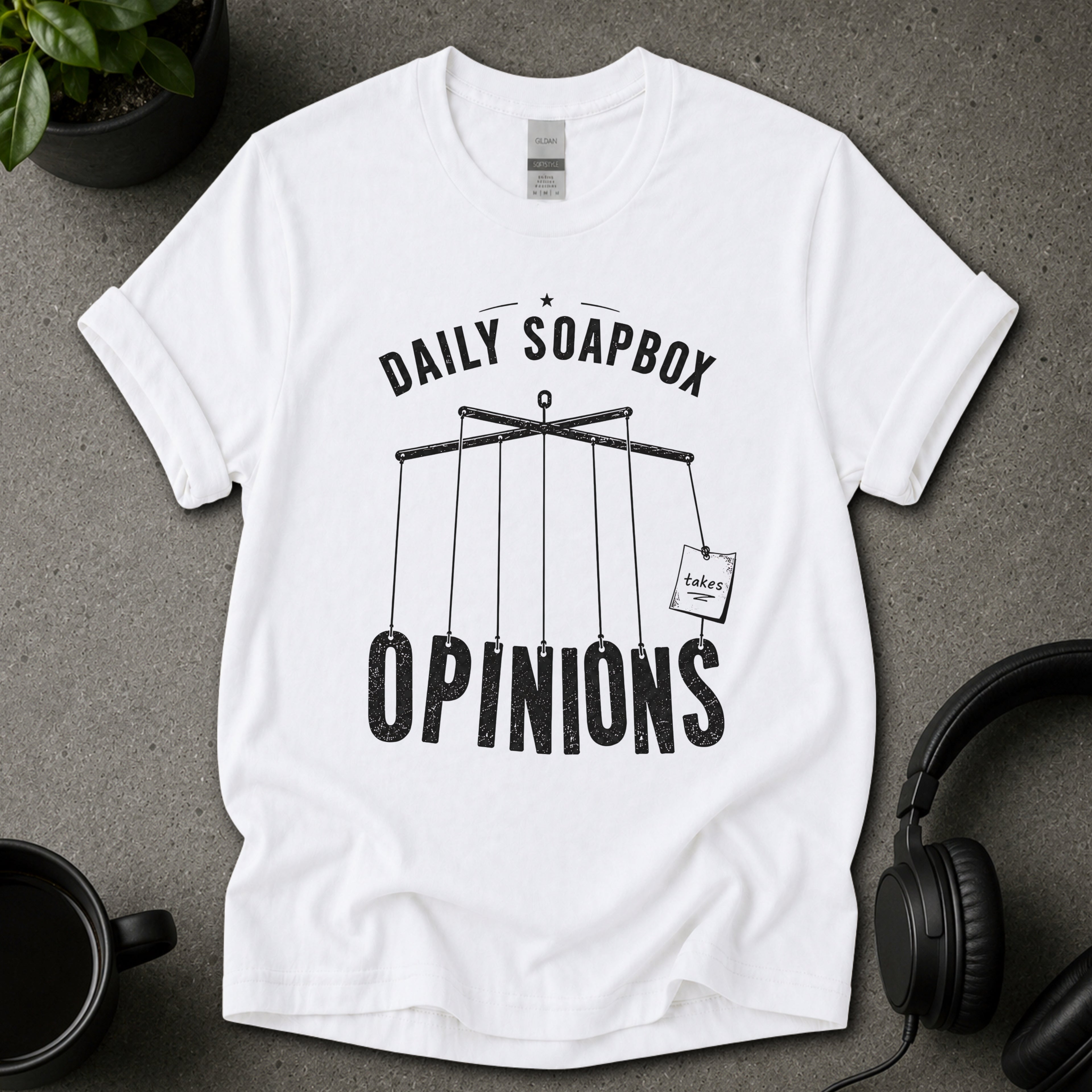 White t-shirt with 'Daily Soapbox Opinions' graphic on a gray surface with a mug and headphones.