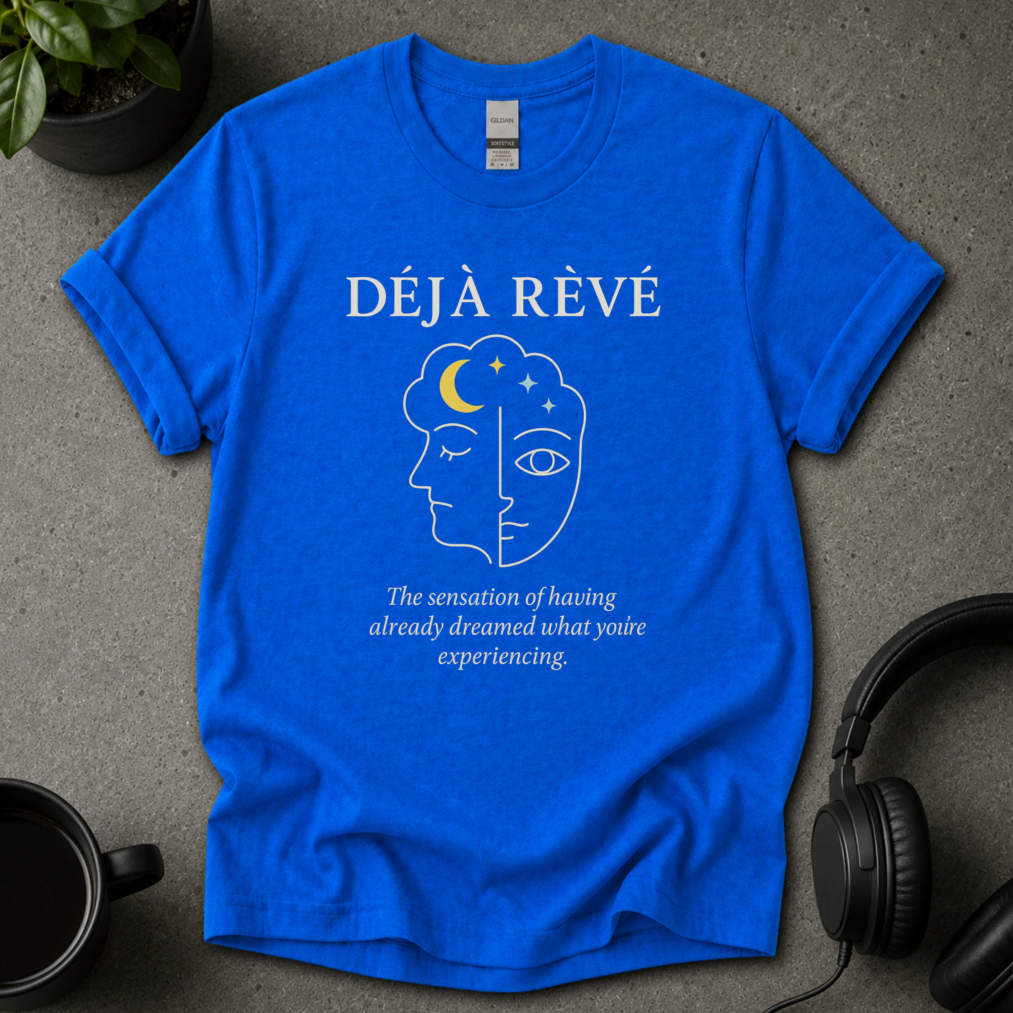 Blue t-shirt with 'Déj  Rèvé' design on a gray surface with headphones and a mug.