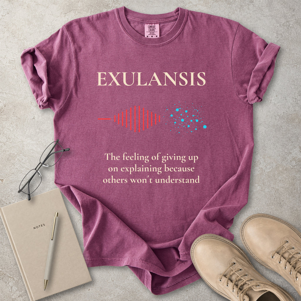 Purple t-shirt with 'EXULANSIS' text and graphics on a concrete surface with glasses, notebook, and shoes.
