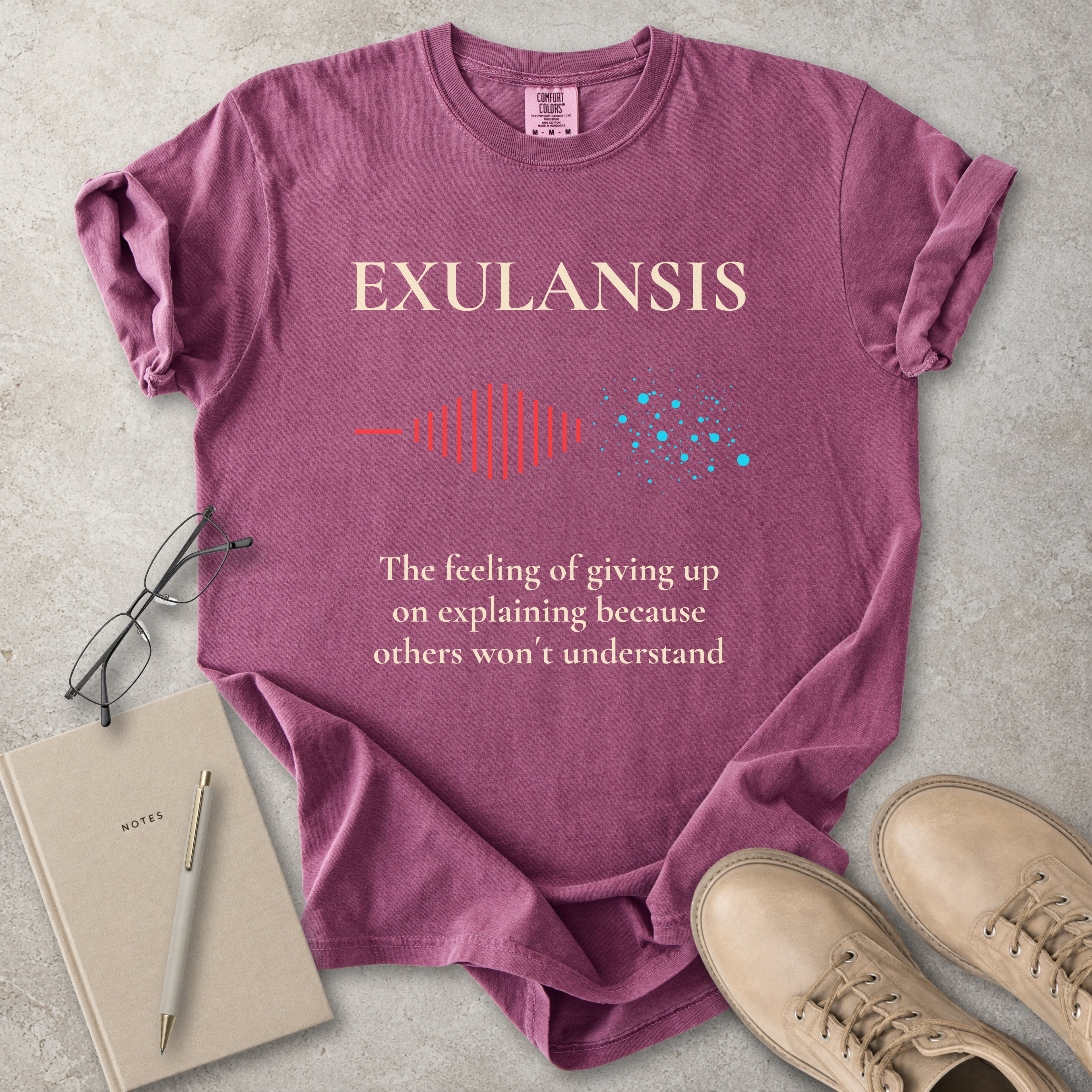Purple t-shirt with 'EXULANSIS' text and graphics on a concrete surface with glasses, notebook, and shoes.