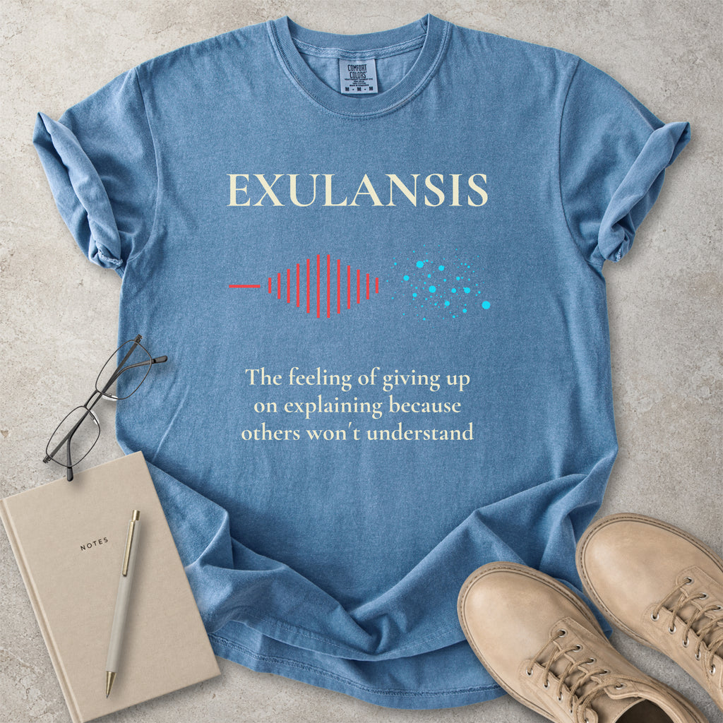 Blue t-shirt with 'EXULANSIS' text and graphic, placed on a concrete surface with a notebook and shoes.