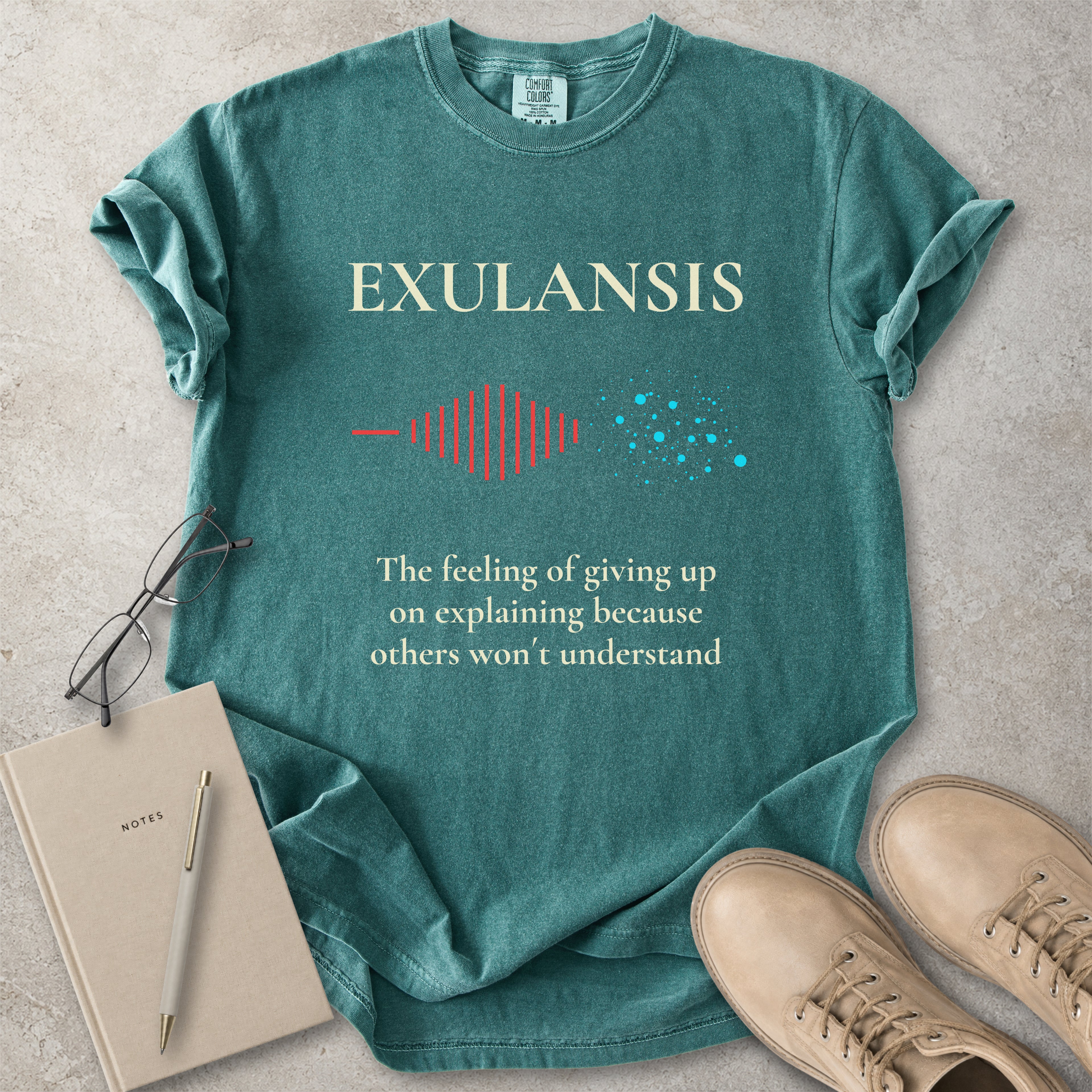 Green t-shirt with 'EXULANSIS' text and graphic, placed on a concrete surface with a notebook and shoes.
