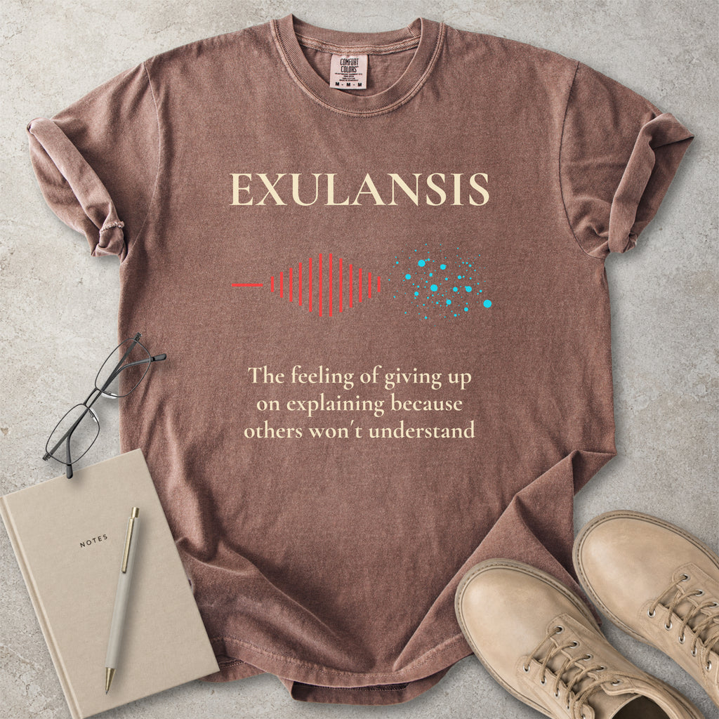 Brown t-shirt with 'EXULANSIS' design and text on a concrete surface with accessories.
