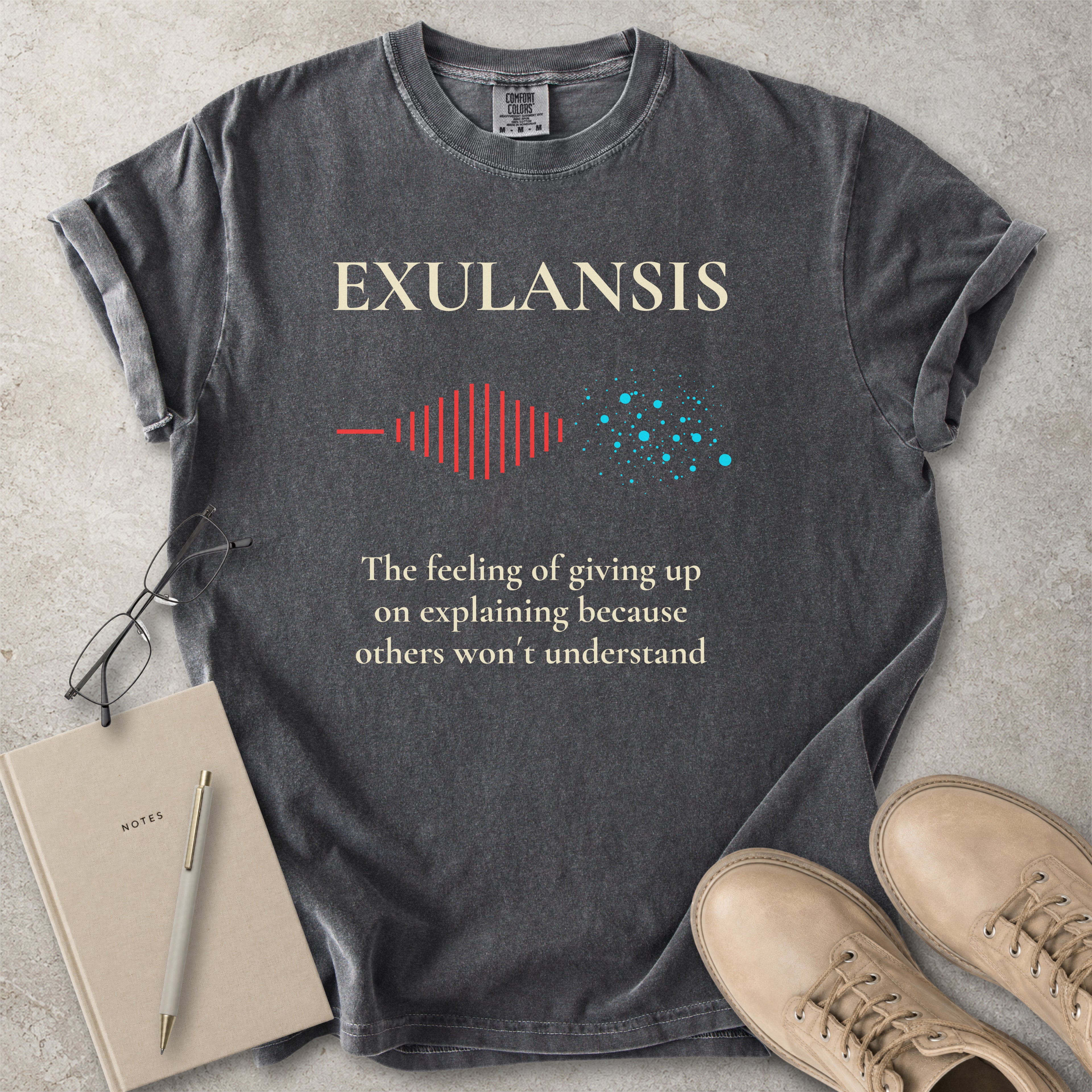 Gray t-shirt with 'Exulansis' design and text on a concrete surface with a notebook and shoes.