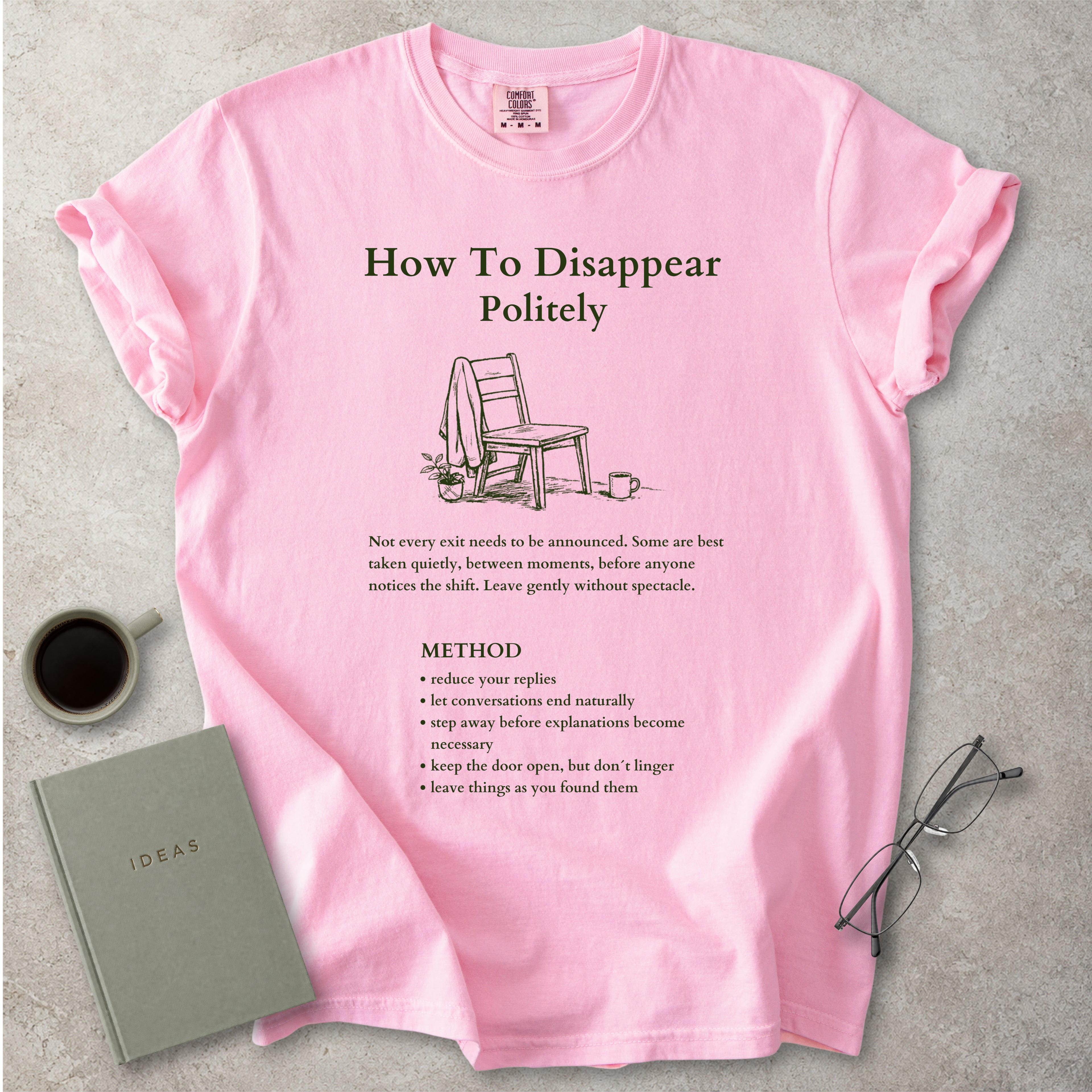 Pink Comfort Colors graphic T-shirt with green How To Disappear Politely design, chair illustration and quiet exit method text, styled with coffee, notebook and glasses