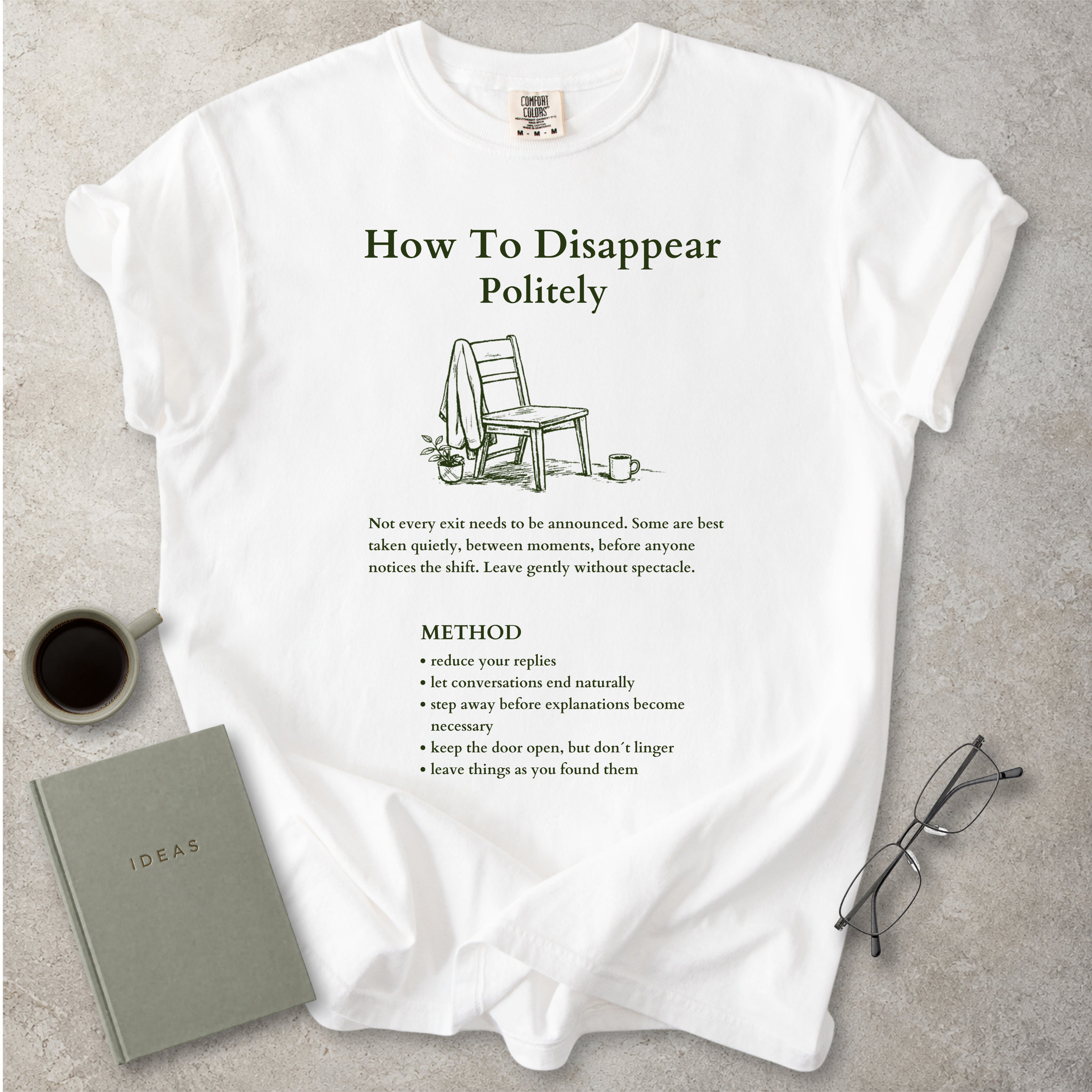 White Comfort Colors graphic T-shirt with green How To Disappear Politely design, chair illustration and quiet exit method text, styled with coffee, notebook and glasses