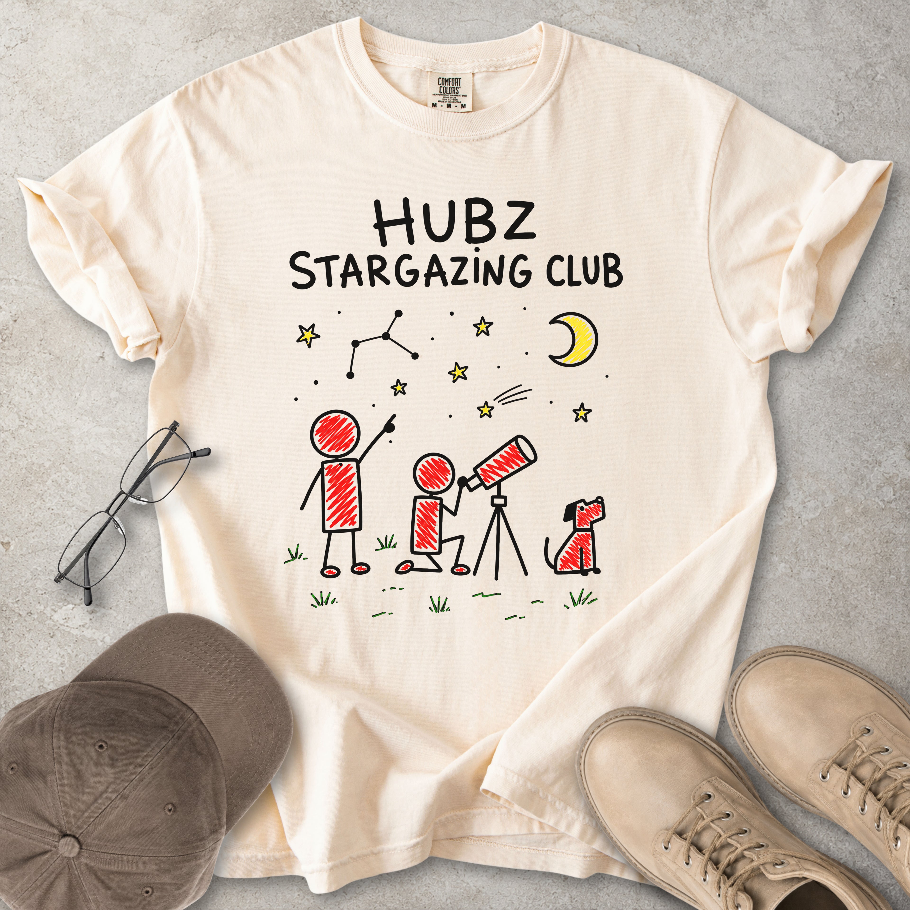 Beige t-shirt with 'HUBZ Stargazing Club' design on a gray surface with glasses, cap, and shoes.