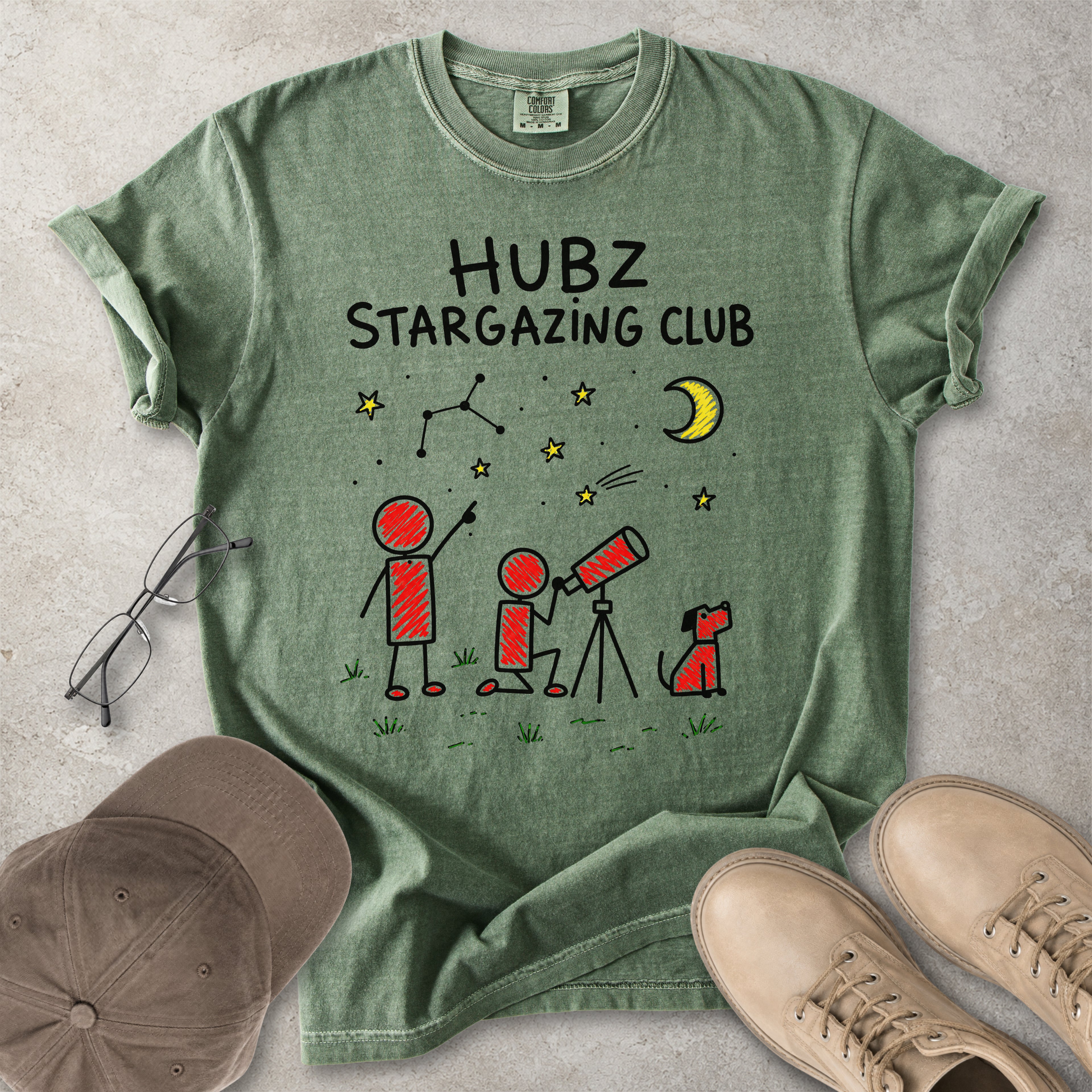 Green t-shirt with 'HUBZ Stargazing Club' design on a concrete surface with glasses, cap, and shoes.