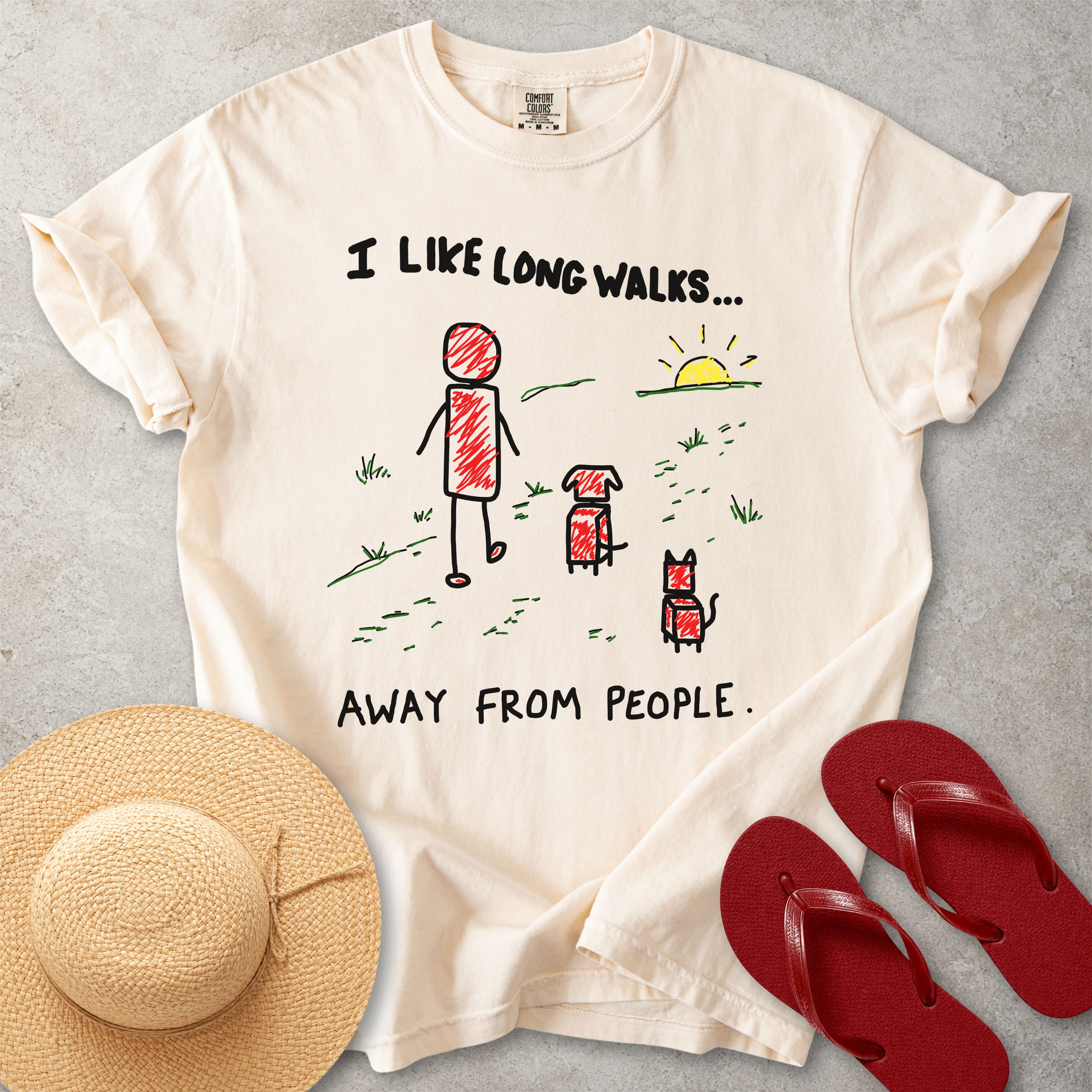 Beige Comfort Colors graphic T-shirt with hand-drawn I like long walks away from people design showing a person walking with a dog and cat, styled with straw hat and red flip flops
