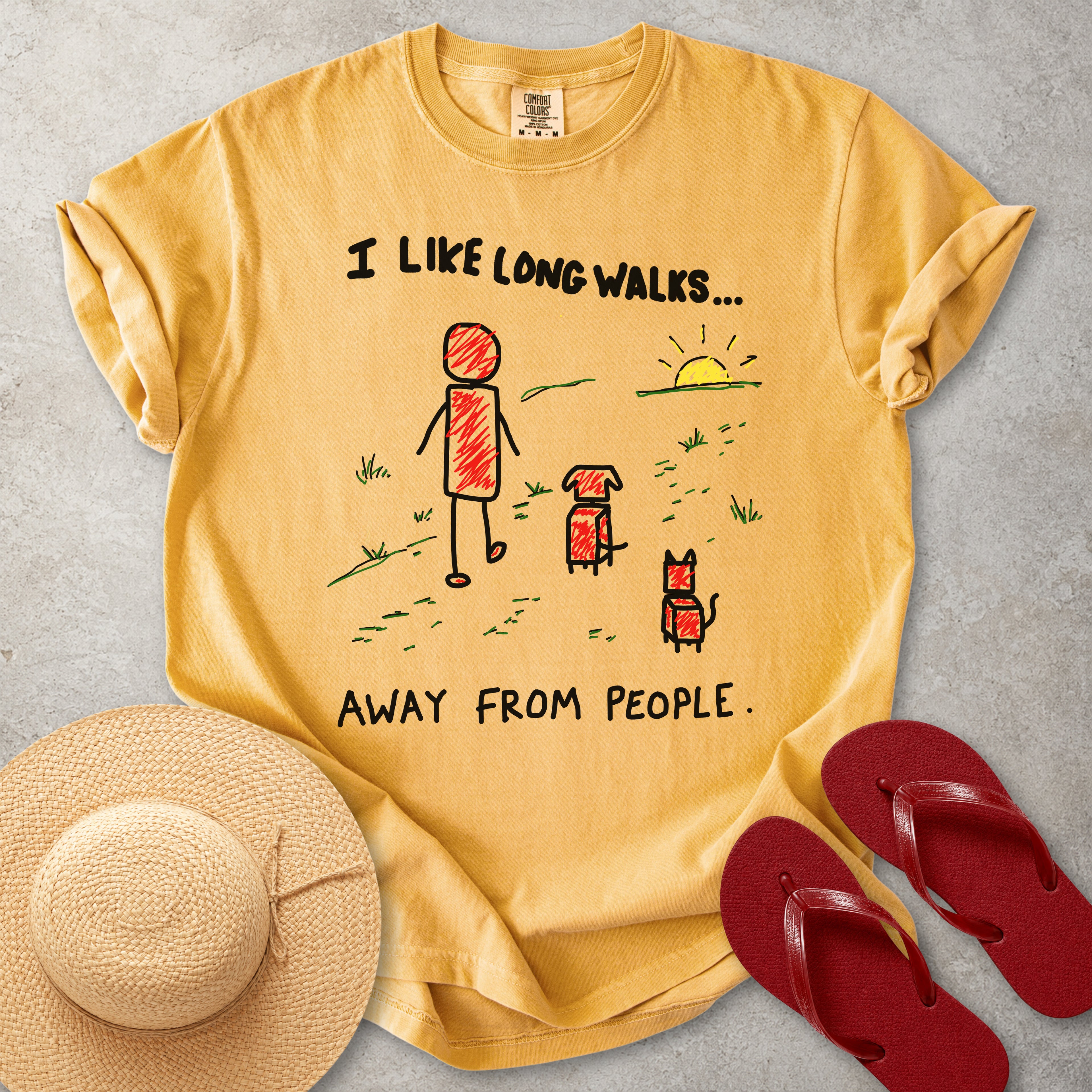 Mustard yellow Comfort Colors graphic T-shirt with hand-drawn I like long walks away from people design showing a person walking with a dog and cat, styled with straw hat and red flip flops