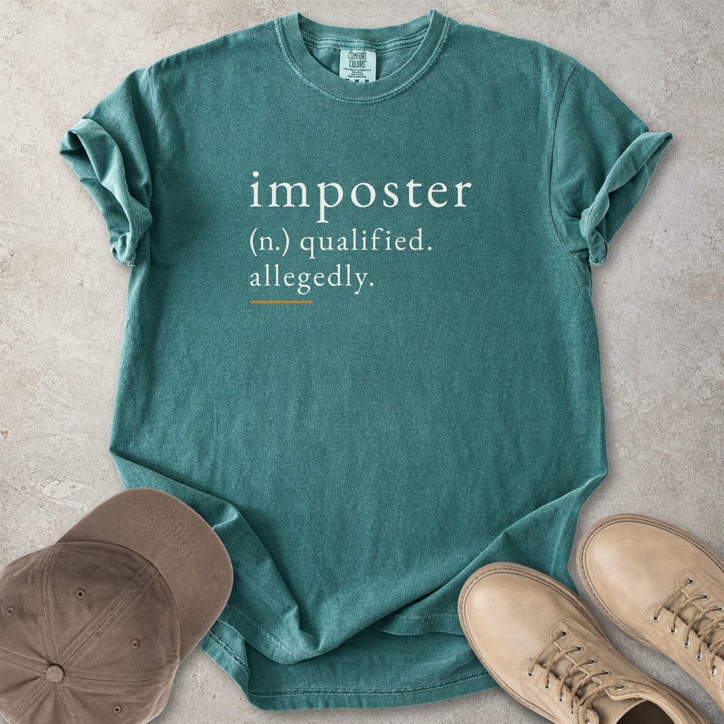 Green t-shirt with 'imposter' definition text on a concrete surface with a cap and shoes.