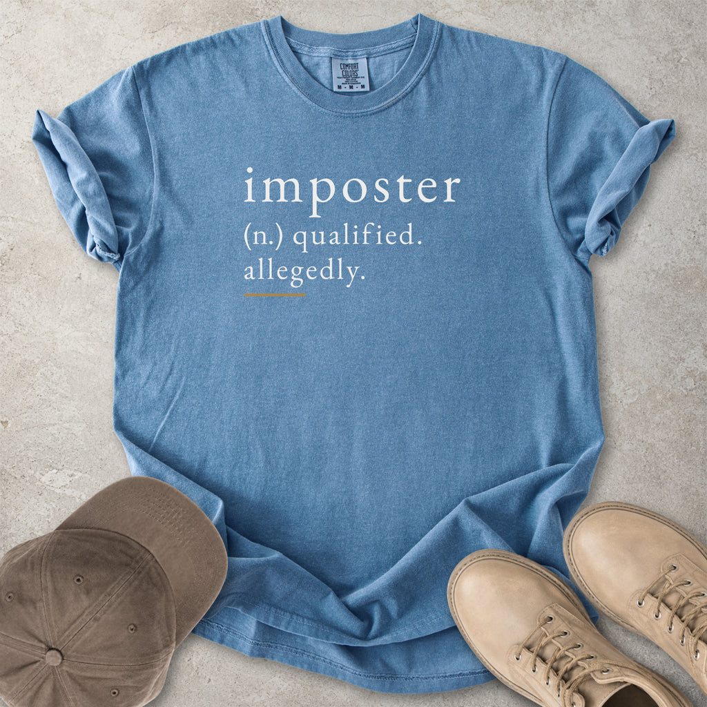 Blue t-shirt with 'imposter' definition text on a concrete surface with shoes and a cap.