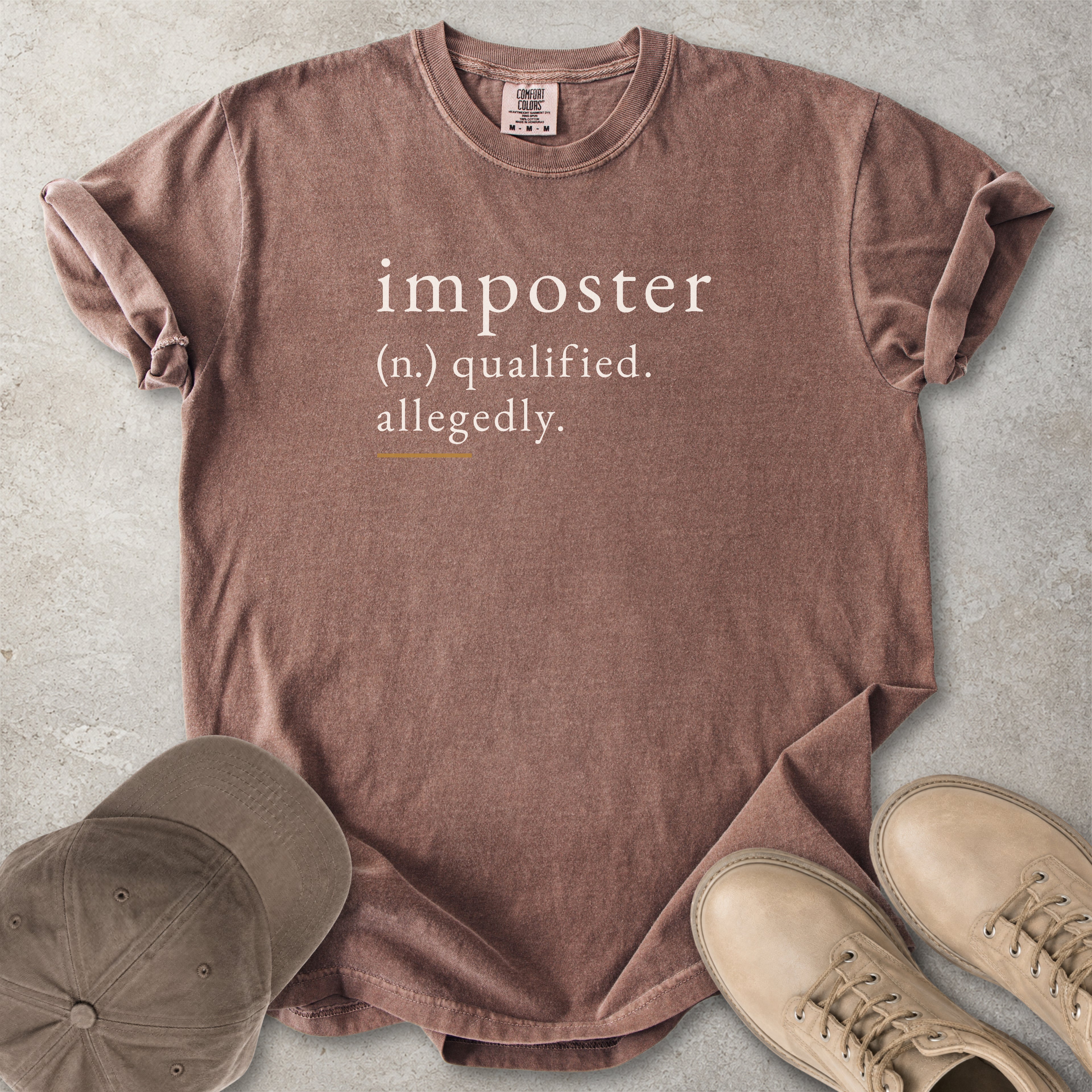 Brown t-shirt with text 'imposter (n.) qualified. allegedly.' on a concrete surface with a cap and shoes.